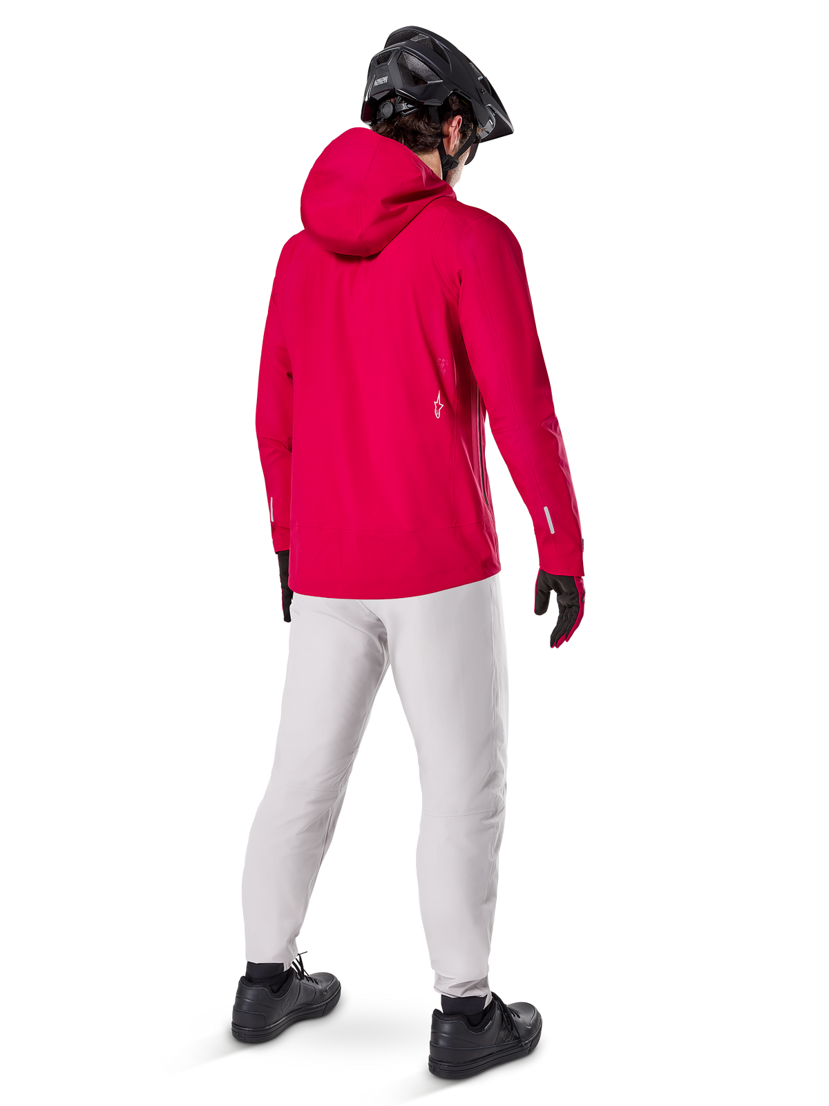 Veste imperméable Alpinestars A-Dura Elite, rouge fluo, vue de dos de trois-quarts, portée avec un pantalon gris clair et un casque de VTT noir, dotée d'une capuche et de détails réfléchissants, veste de pluie technique haute performance pour le trail