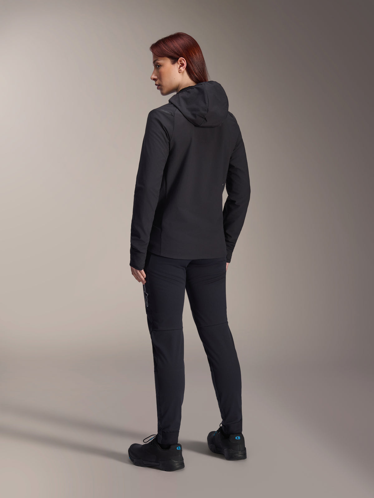 Veste thermique Alpinestars Stella A-Dura, veste de VTT pour femme, noire, vue de dos mettant en avant la capuche isolée et la coupe régulière, portée avec un pantalon et des chaussures noirs assortis, isolation thermique légère pour rouler par temps froid