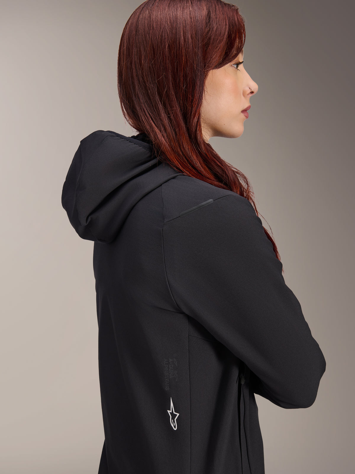Veste thermique Alpinestars Stella A-Dura, veste de cyclisme pour femme, noire, profil latéral d'une femme aux cheveux roux portant la veste, dotée d'une capuche intégrée et d'un logo étoile Alpinestars blanc sur le côté, équipement thermique isolant léger pour le VTT