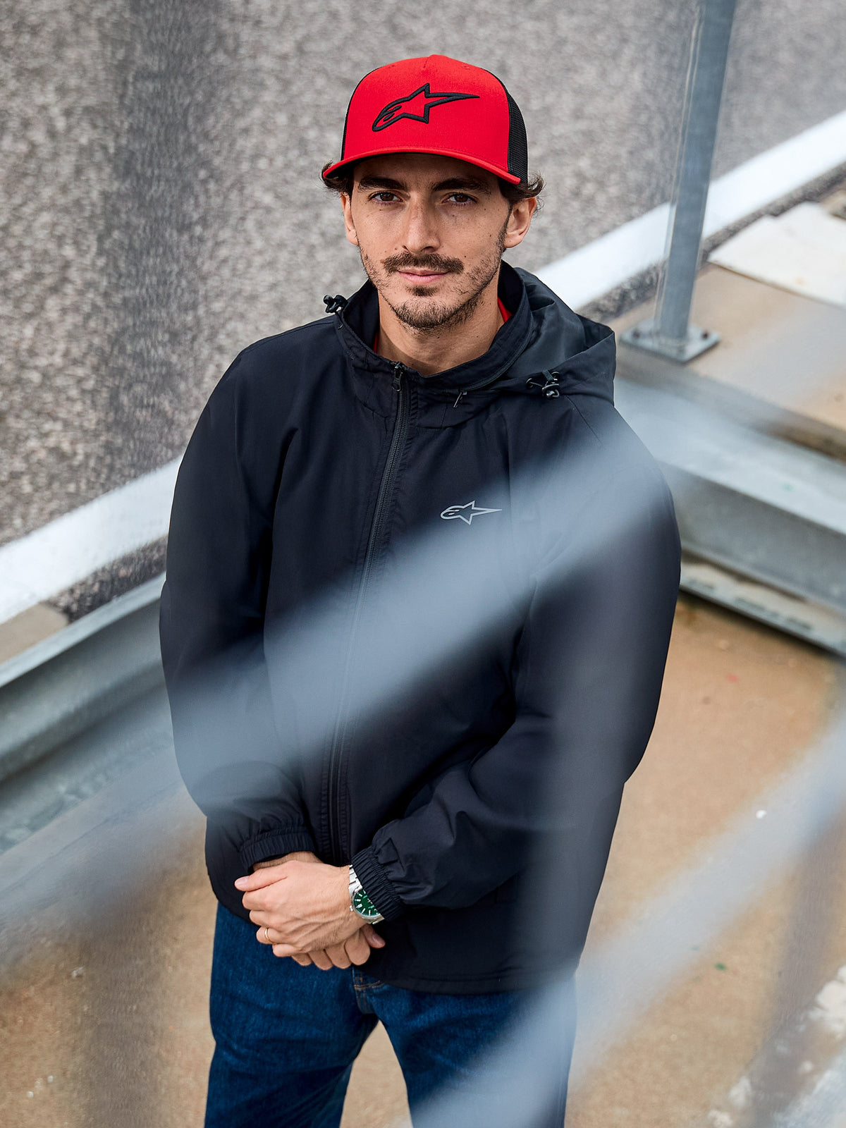 Homme portant la veste Alpinestars Overhaul Racer noire, une veste de pluie à capuche résistante à l'eau avec un petit logo sur la poitrine, accompagnée d'une casquette Alpinestars rouge, vêtement lifestyle motorsport décontracté