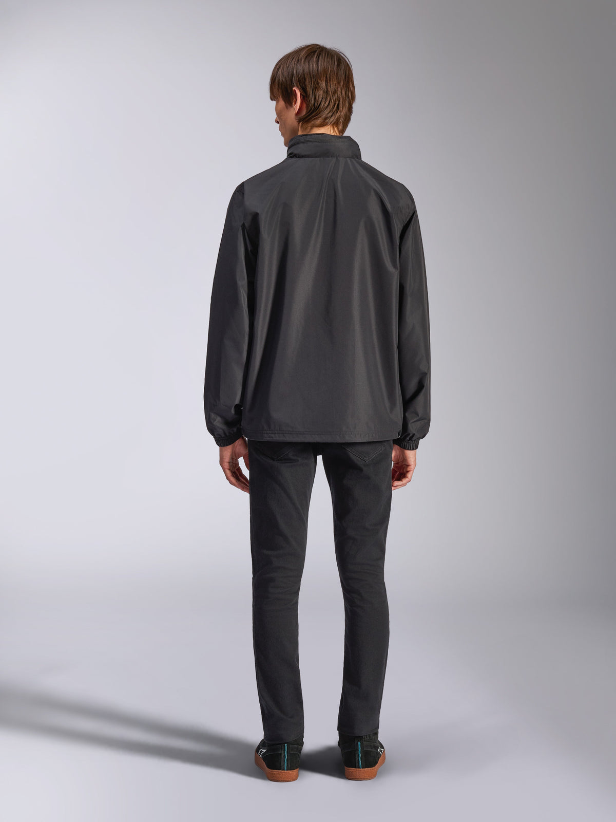 Veste Alpinestars Overhaul Racer, noir, vue de dos d'un homme portant la veste de pluie, tissu résistant à l'eau 2.5L, poignets élastiqués et capuche fixe, associée à un pantalon noir, vêtement d'extérieur décontracté inspiré du sport automobile
