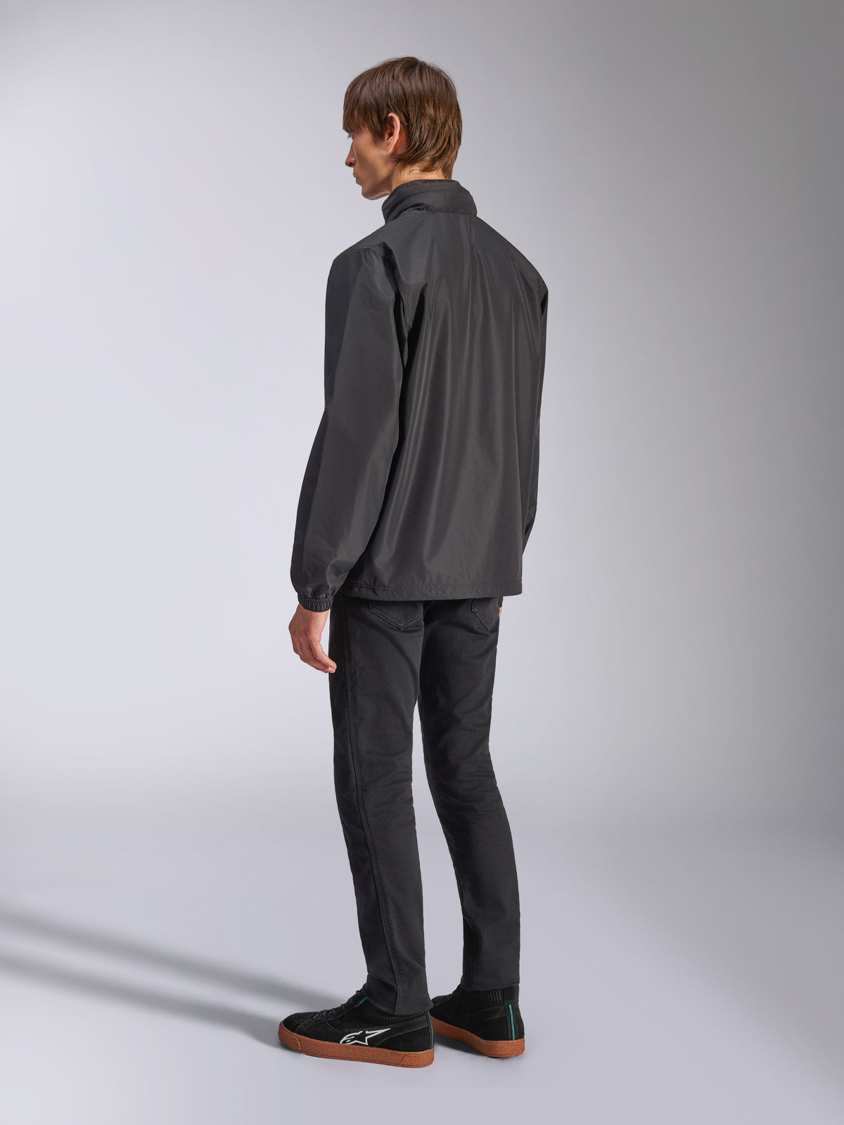 Veste Alpinestars Overhaul Racer, noire, vue de dos aux trois-quarts, portée par un mannequin avec un jean noir et des baskets noires, veste de pluie légère résistante à l'eau pour un style de vie décontracté inspiré du sport automobile