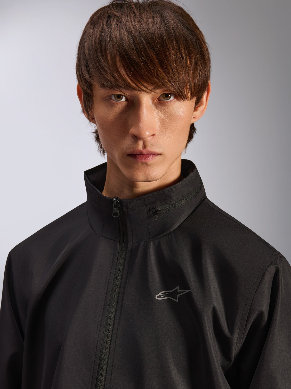 Veste Alpinestars Overhaul Racer, veste de pluie noire, portrait rapproché d'un mannequin portant la veste, avec un col montant et un petit logo Astars argenté sur la poitrine, vêtement d'extérieur décontracté d'inspiration sport mécanique