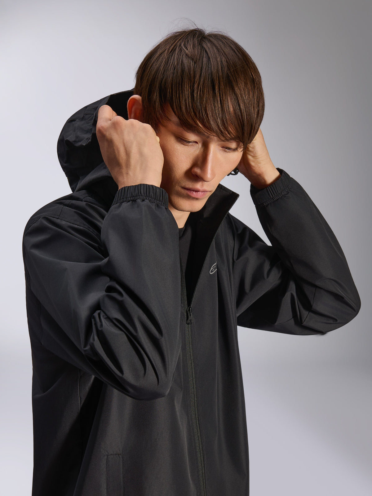 Veste Alpinestars Overhaul Racer, veste de pluie à capuche noire, portée par un mannequin avec la capuche relevée, dotée d'un tissu 2,5L résistant à l'eau, poignets élastiqués et logo minimaliste sur la poitrine, vêtement d'extérieur décontracté inspiré du sport automobile