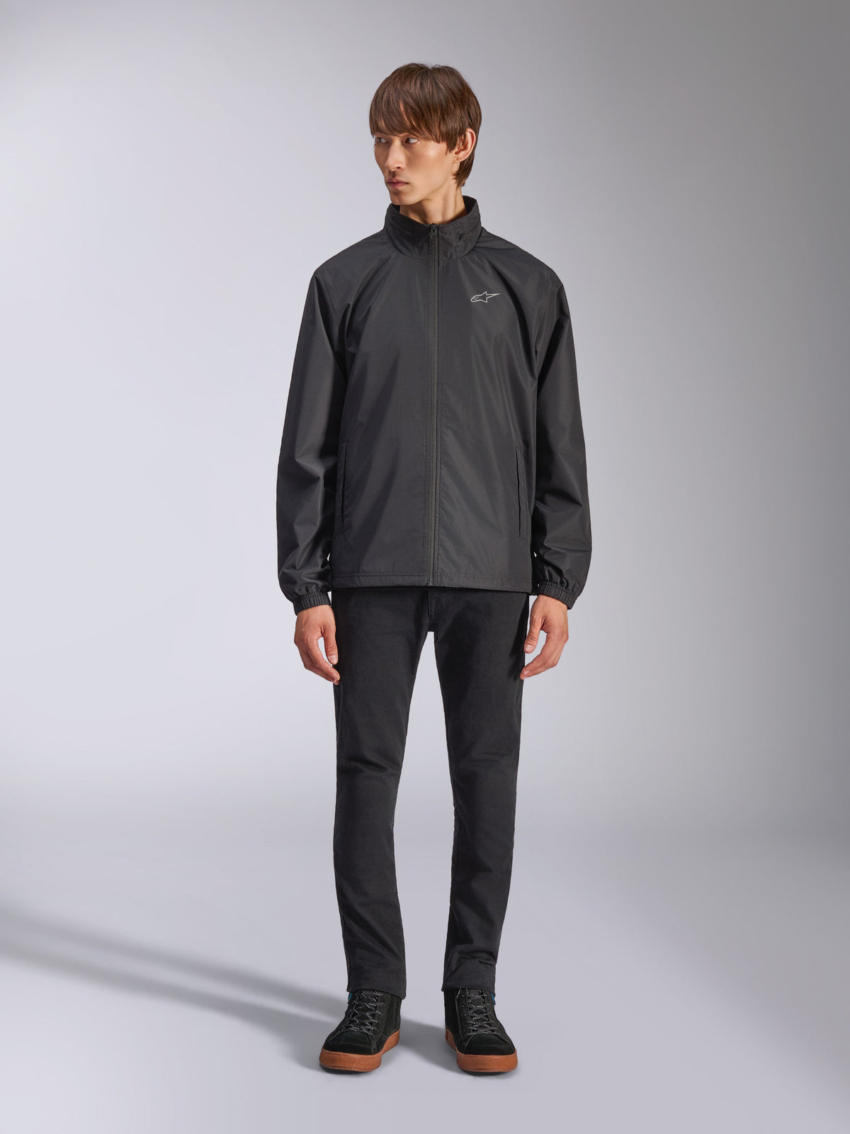 Veste Alpinestars Overhaul Racer, veste de pluie noire pour homme, vue en pied sur un mannequin avec pantalon noir et baskets décontractées, dotée d'un petit logo blanc sur la poitrine, d'un tissu résistant à l'eau et d'un design décontracté inspiré du sport automobile
