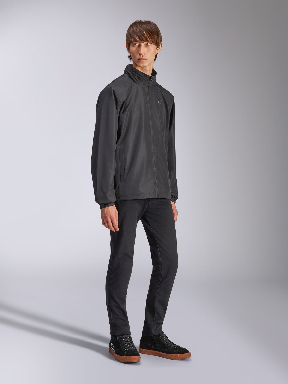 Veste Alpinestars Overhaul Racer, veste de pluie noire, portée avec un pantalon noir et des baskets noires à semelles en gomme, logo étoile blanc sur la poitrine, fermeture éclair à l'avant, vêtement de sport décontracté d'inspiration sport automobile