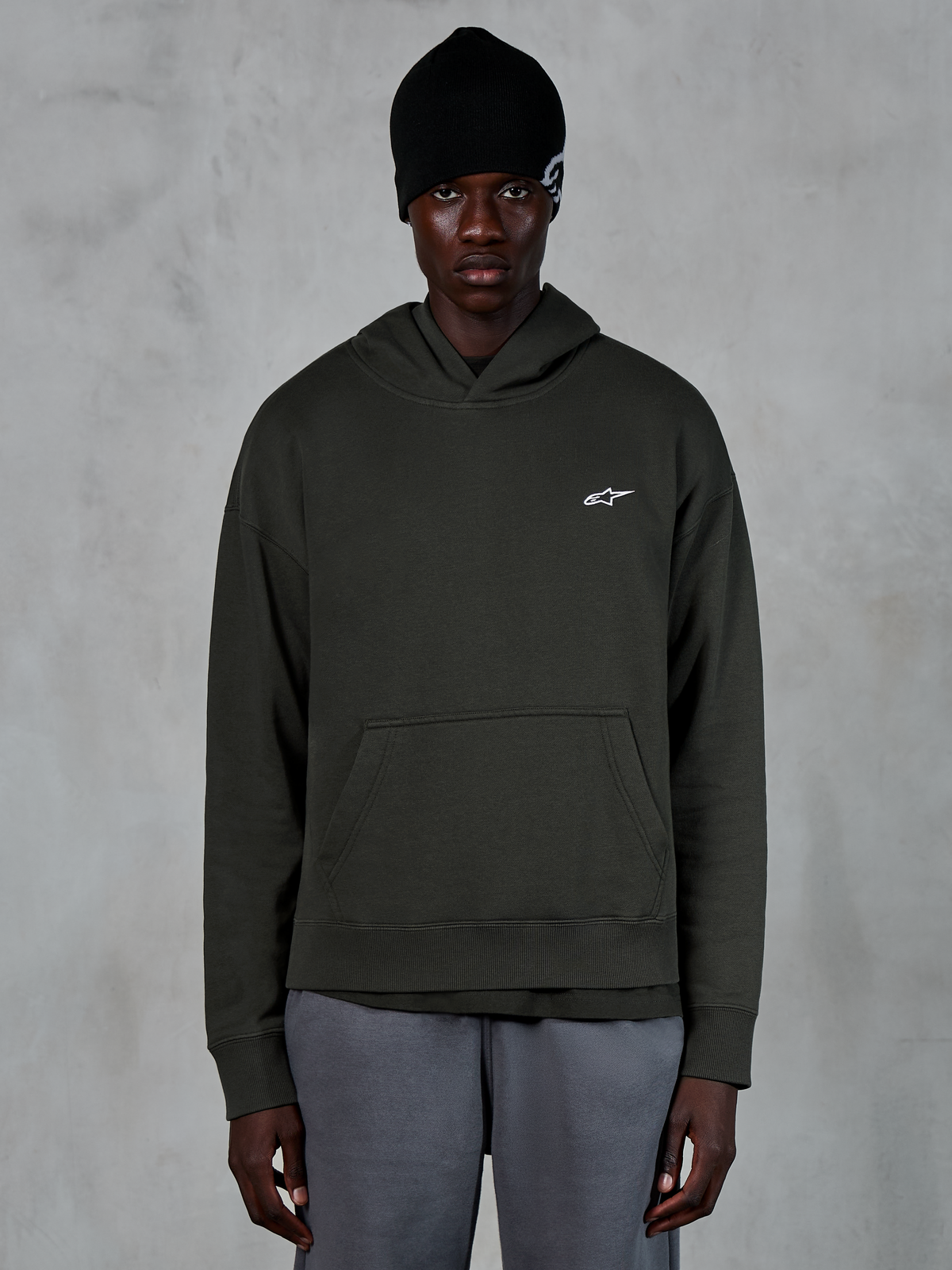 Sweat à capuche Alpinestars Essentials Ageless, style streetwear décontracté, pullover Off Black avec un petit logo Alpinestars blanc sur la poitrine, porté avec un bonnet noir, design minimaliste avec une poche kangourou à l'avant et finition délavée