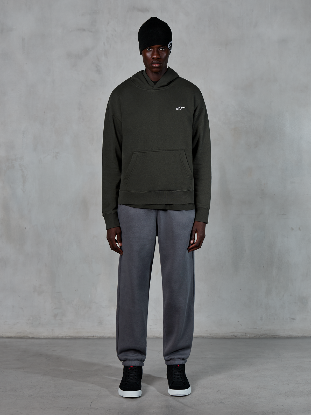 Modèle portant le sweat à capuche Alpinestars Essentials Ageless, streetwear décontracté, Off Black avec un petit logo blanc sur la poitrine, associé à un jogging gris, un bonnet noir et des baskets noires, design minimaliste inspiré du sport automobile