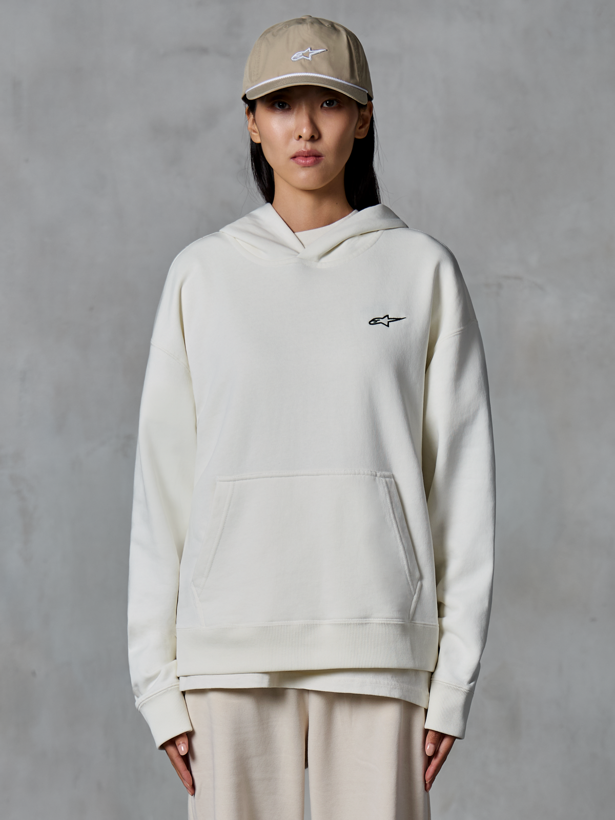 Sweat à capuche Alpinestars Essentials Ageless, sweat à capuche en molleton blanc cassé, mannequin femme portant une casquette beige et un jogging crème assorti, petit logo Alpinestars noir sur la poitrine, poche kangourou à l'avant, coupe décontractée, streetwear décontracté d'inspiration sport automobile