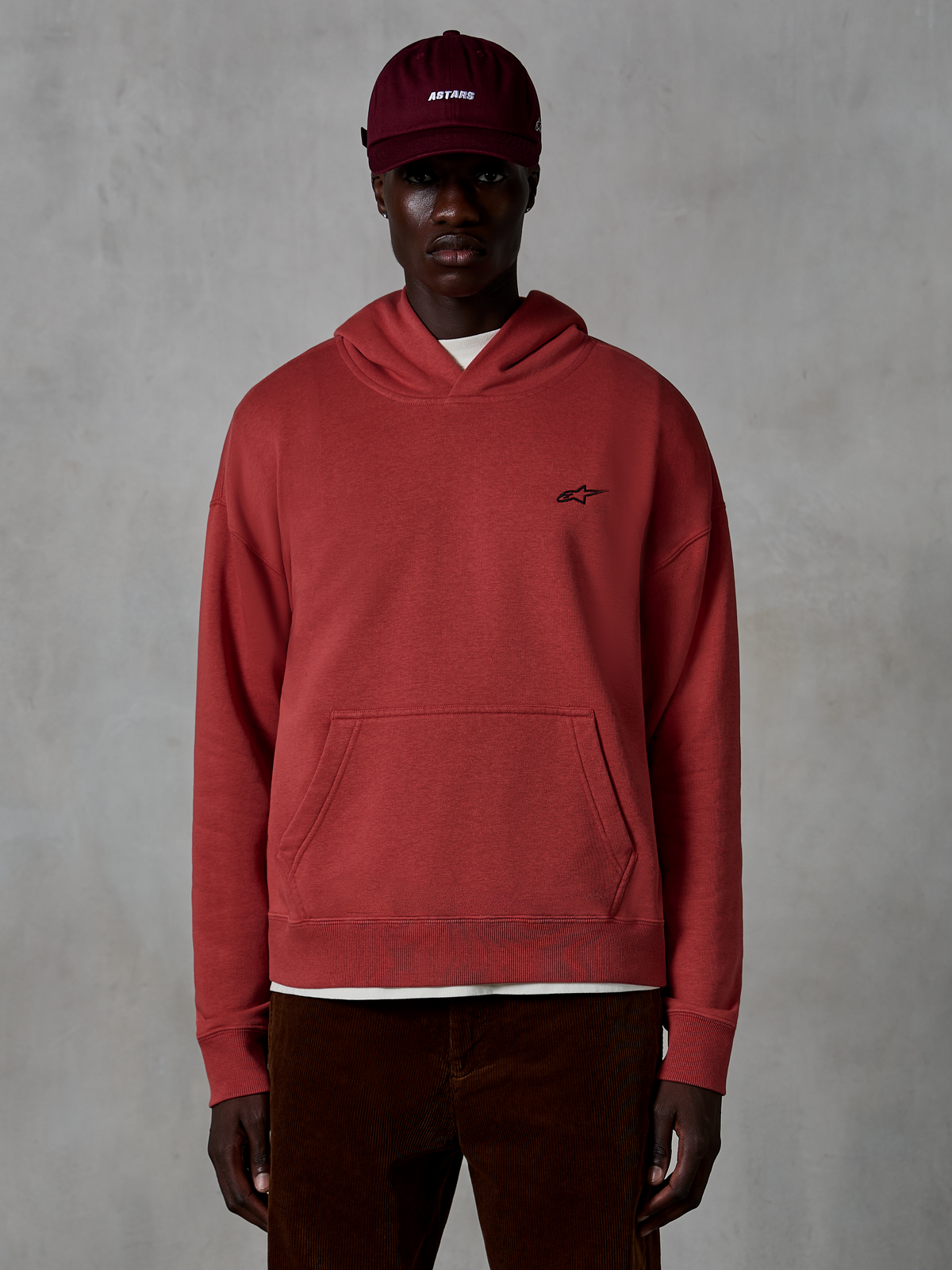 Sweat à capuche Alpinestars Essentials Ageless, style streetwear décontracté, couleur rouge brique, avec un petit logo étoile noir sur la poitrine, poche kangourou, finition délavée, porté par un mannequin avec une casquette bordeaux et un pantalon en velours côtelé marron
