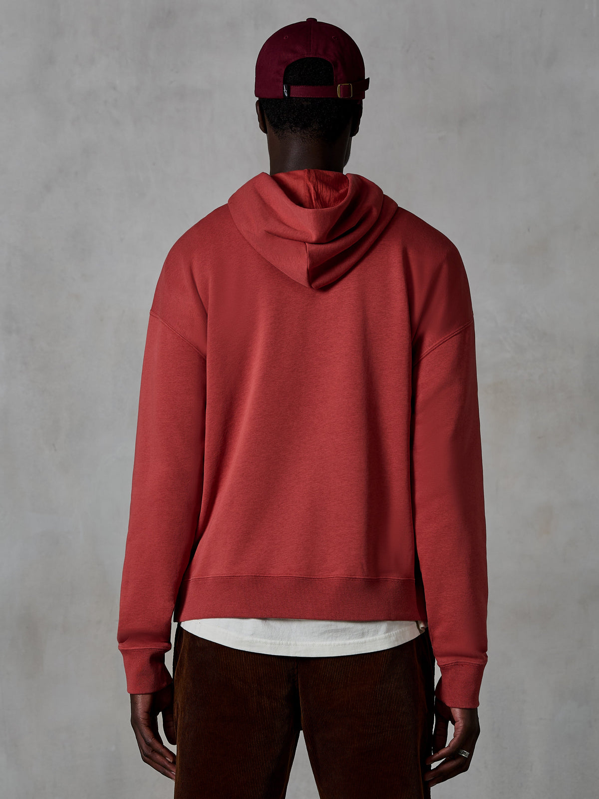 Sweat à capuche Alpinestars Essentials Ageless, sweat à capuche décontracté en molleton, rouge brique, vue de dos d'un mannequin portant des vêtements lifestyle, présentant un design épuré avec ourlet et poignets côtelés, porté avec un pantalon en velours côtelé marron et une casquette bordeaux