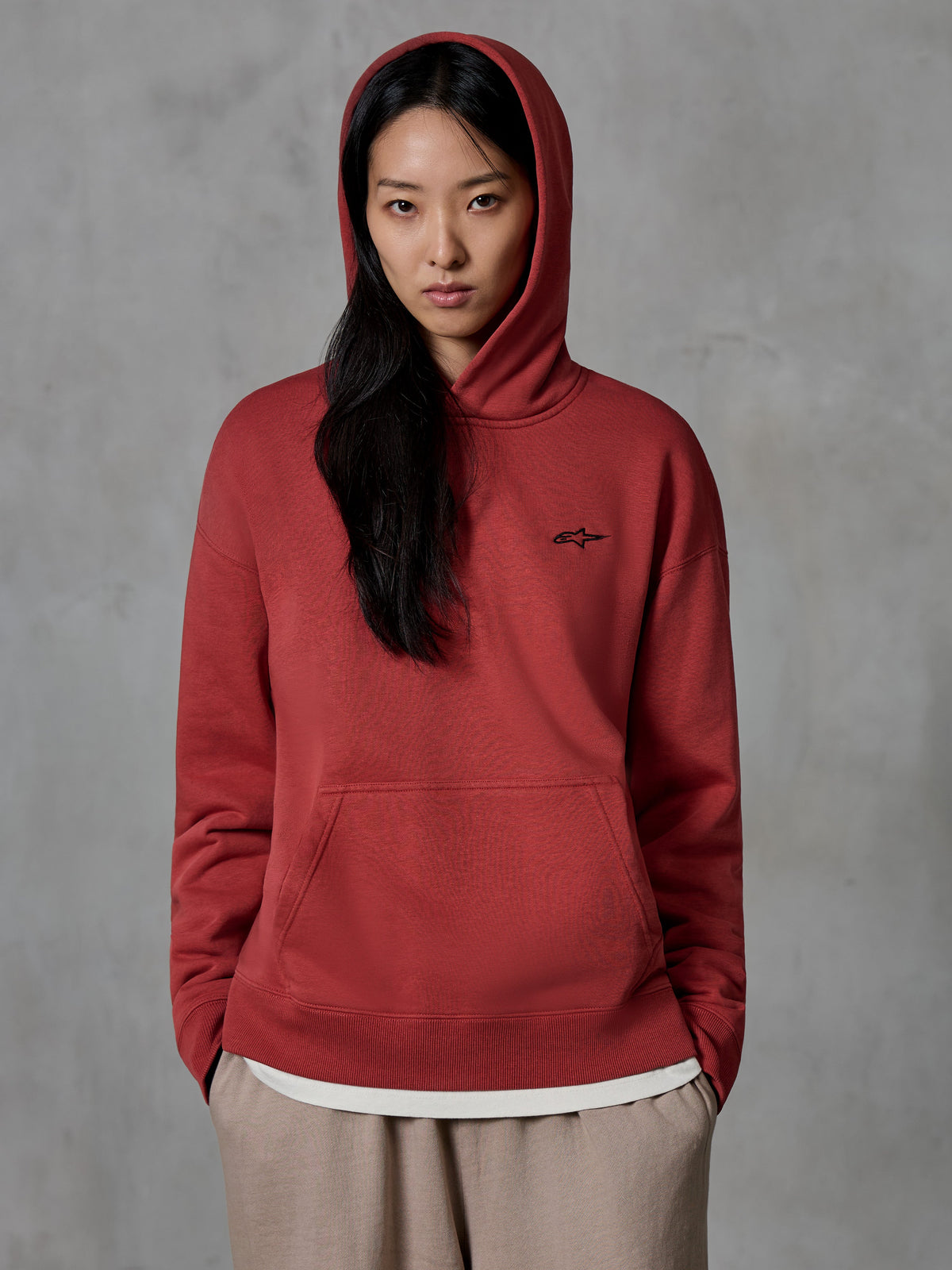 Sweat à capuche Alpinestars Essentials Ageless, sweat rouge brique porté par une mannequin avec la capuche relevée, petit logo étoile noir brodé sur la poitrine, poche kangourou, streetwear décontracté inspiré du sport automobile.