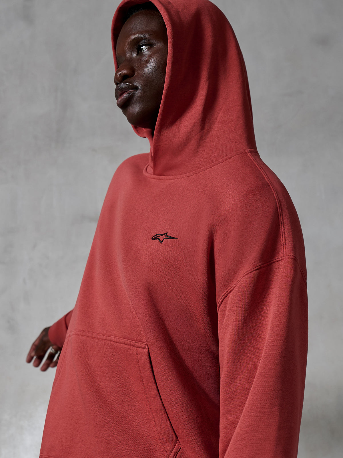 Sweat à capuche Alpinestars Essentials Ageless, sportswear décontracté, rouge brique, porté par un mannequin avec la capuche relevée, petit logo noir brodé, texture en molleton doux, coupe décontractée style streetwear