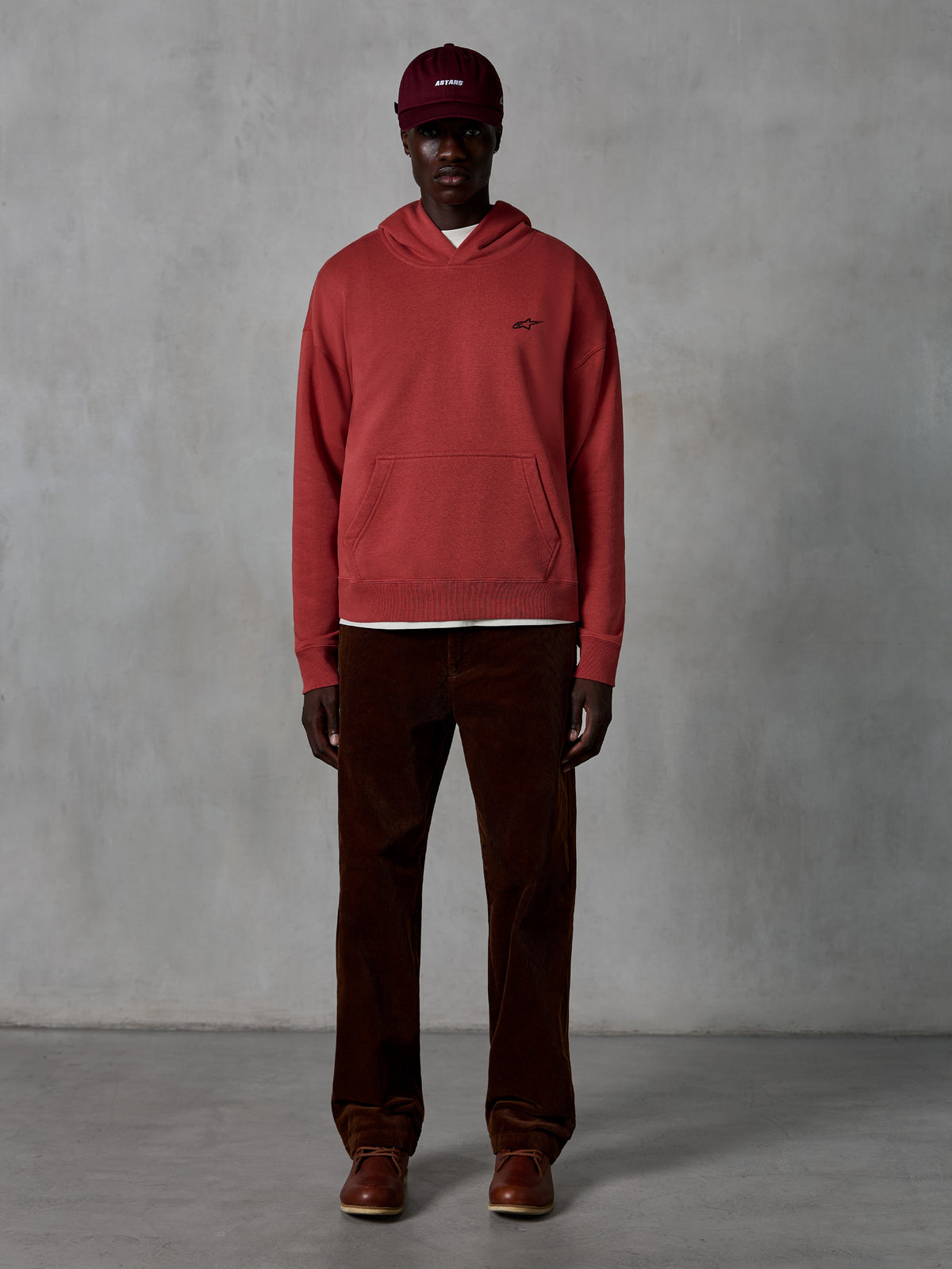 Sweat à capuche Essentials Ageless Alpinestars, sweat à capuche sportswear décontracté rouge brique avec finition délavée à la pierre, arborant un petit logo Alpinestars noir sur la poitrine et une poche kangourou à l'avant, porté par un mannequin avec une casquette bordeaux et un pantalon en velours côtelé marron pour un look streetwear épuré.
