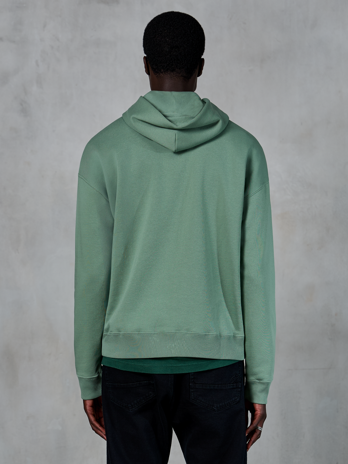 Sweat à capuche Alpinestars Essentials Ageless, sportswear décontracté, vert Cactus, vue de dos sur modèle, finition lavage à la pierre, porté avec un pantalon noir, design lifestyle minimaliste