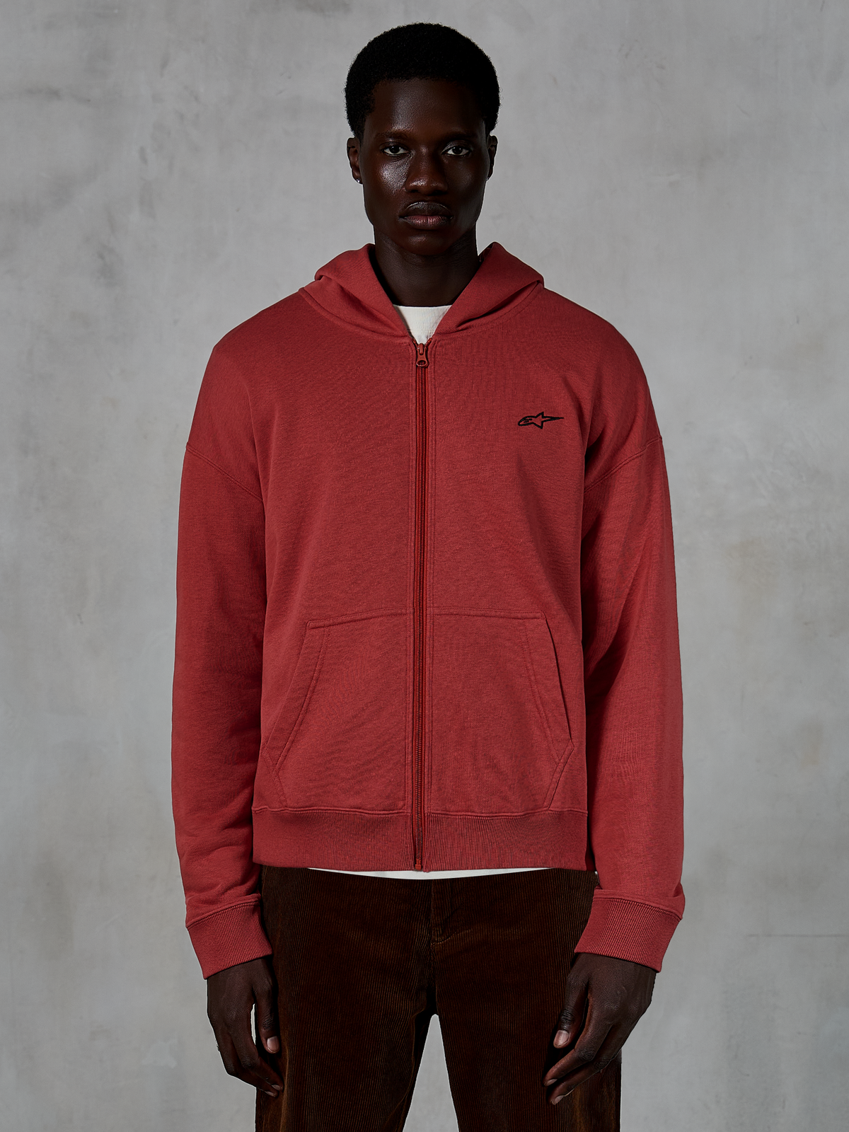 Alpinestars Essentials Ageless Zip Hoodie, sweat à capuche entièrement zippé rouge brique, porté par un mannequin avec un pantalon en velours côtelé marron, arborant un petit logo noir sur la poitrine, coupe décontractée style streetwear