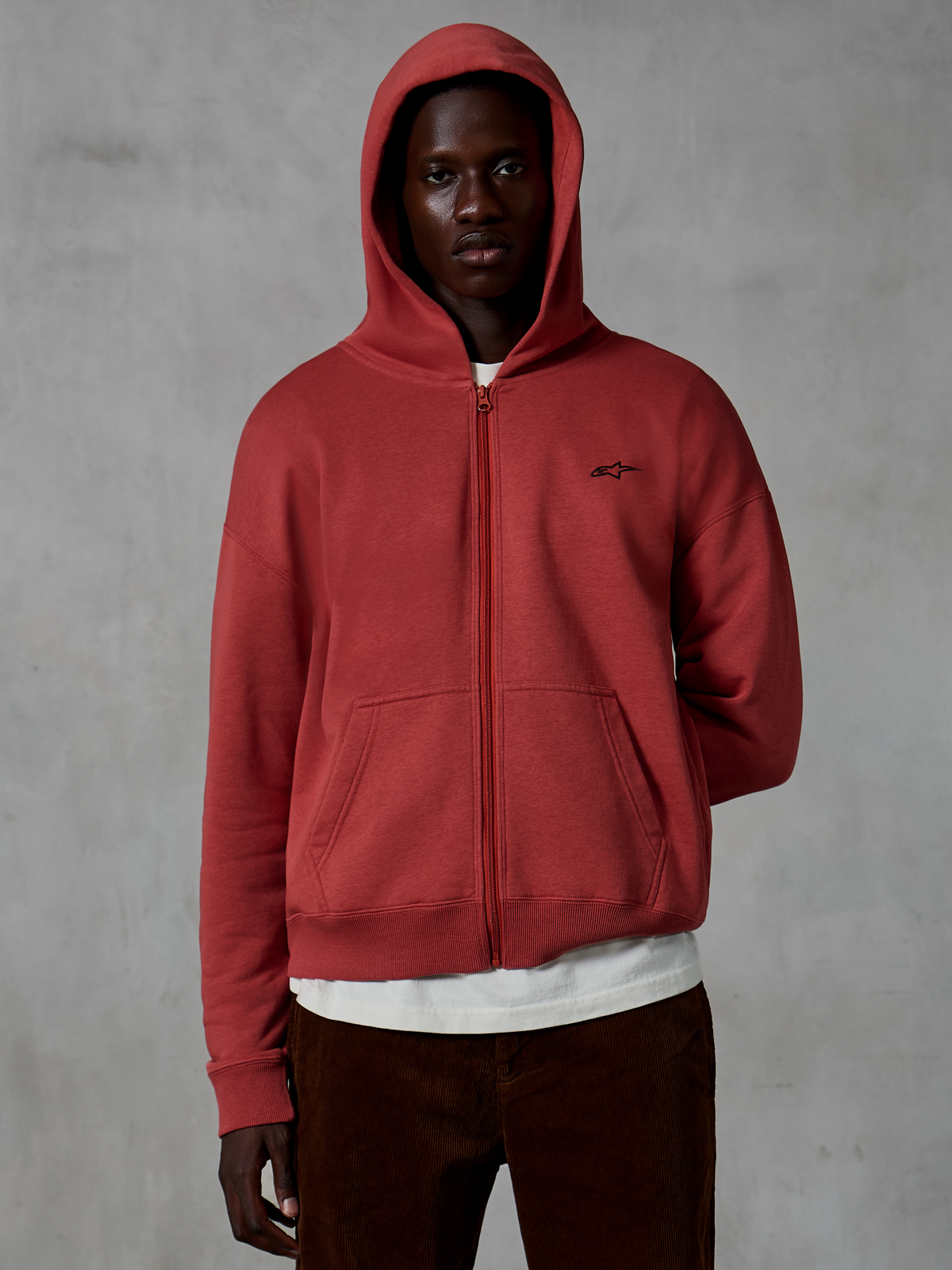 Alpinestars Essentials Ageless Zip Hoodie, sweat à capuche zippé sportswear en rouge brique, modèle à fermeture éclair avec capuche relevée, petit logo noir sur la poitrine, porté par un mannequin avec un t-shirt blanc et un pantalon en velours côtelé marron sur un fond gris.
