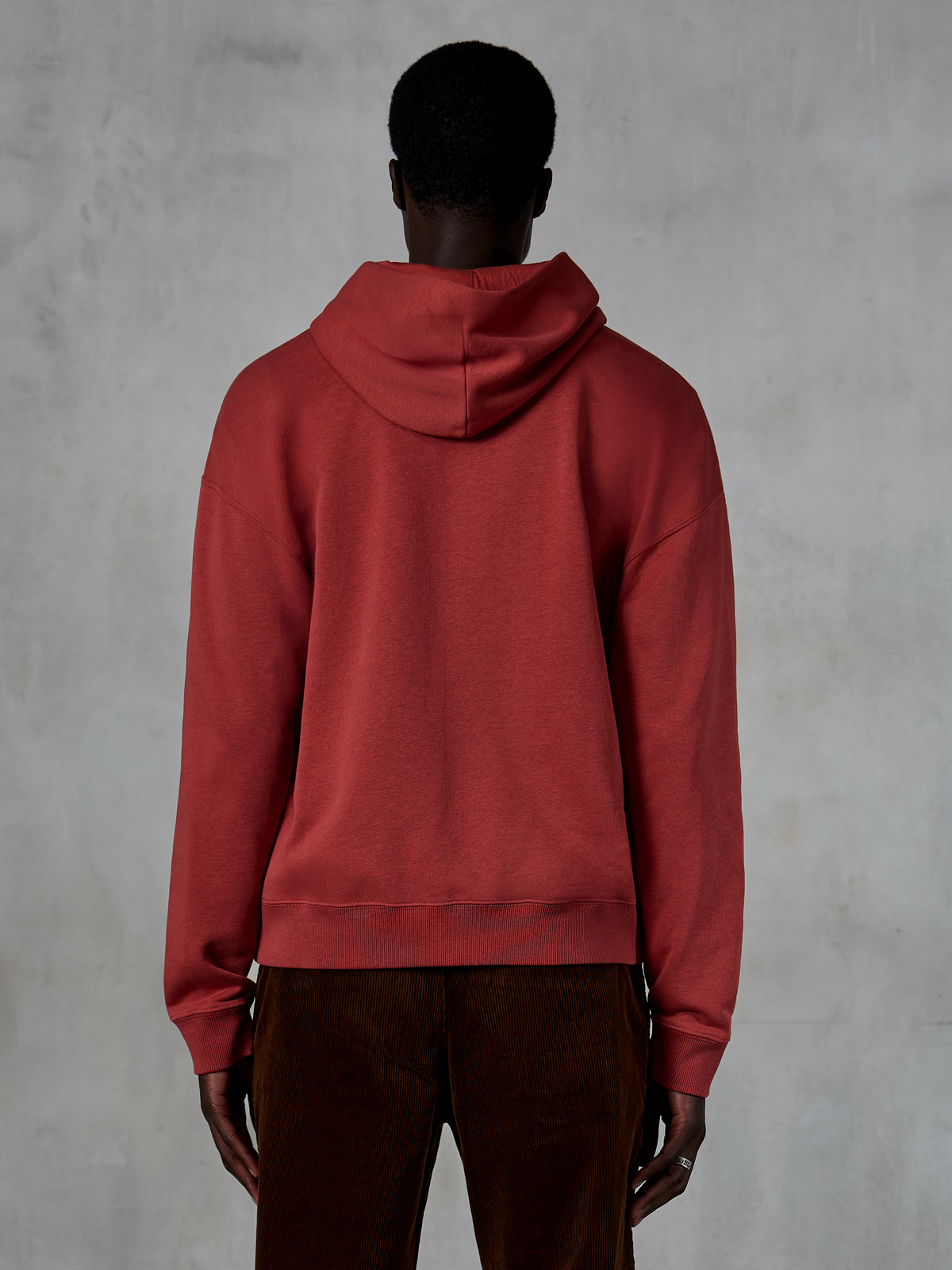 Alpinestars Essentials Ageless Zip Hoodie, rouge brique, vue de dos montrant une coupe décontractée et une capuche avec cordon de serrage, porté avec un pantalon en velours côtelé marron, streetwear décontracté inspiré du sport automobile