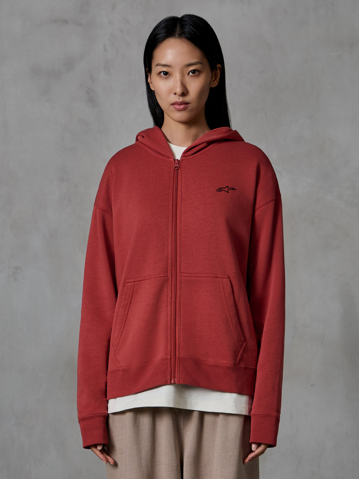 Sweat à capuche zippé Alpinestars Essentials Ageless, rouge brique, style streetwear décontracté, porté par un mannequin, modèle entièrement zippé avec capuche, petit logo Alpinestars noir sur la poitrine, polaire compacte à coupe décontractée.