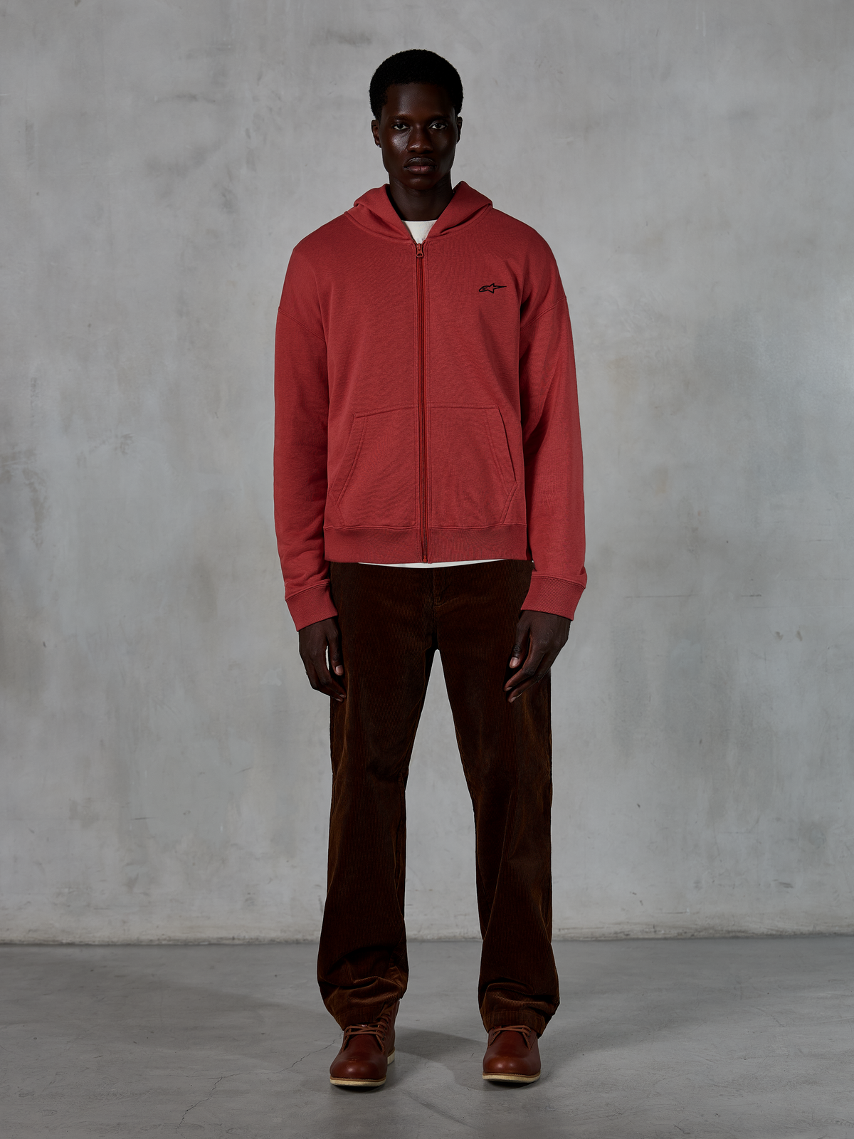 Alpinestars Essentials Ageless Zip Hoodie, streetwear décontracté, rouge brique avec petit logo étoile noir, fermeture éclair intégrale sur le devant, porté par un mannequin avec un pantalon en velours côtelé marron et des bottes, design haut de gamme inspiré du sport automobile