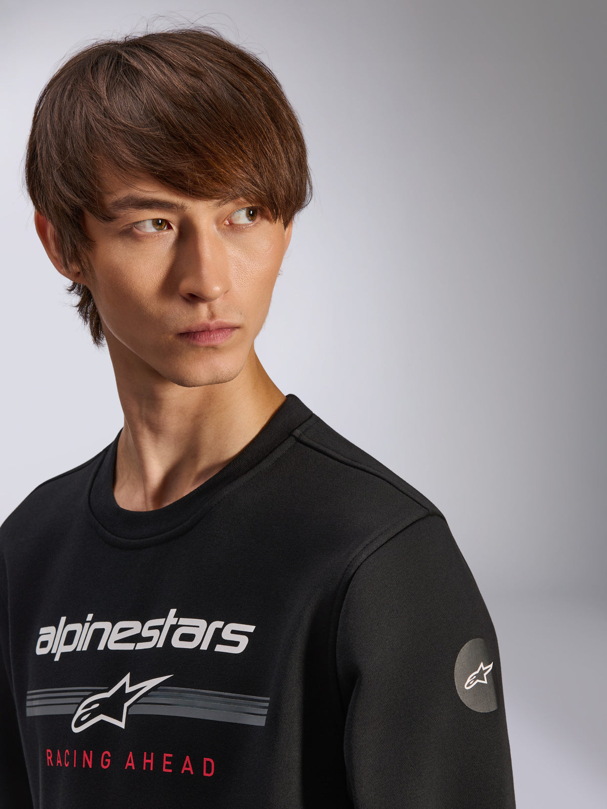 Sweatshirt Alpinestars Bettern Crew, noir, arborant le logo Alpinestars blanc avec des bandes grises et le texte 'RACING AHEAD' en rouge, logo étoile circulaire sur la manche, streetwear décontracté inspiré du sport automobile