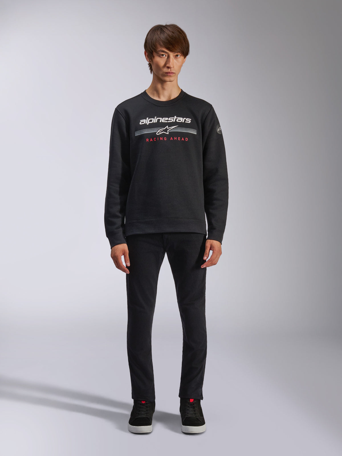Alpinestars Bettern Crew, sweatshirt noir à col rond, logo Alpinestars blanc avec rayures grises horizontales et texte 'RACING AHEAD' rouge, style streetwear décontracté, porté par un mannequin avec pantalon noir et baskets