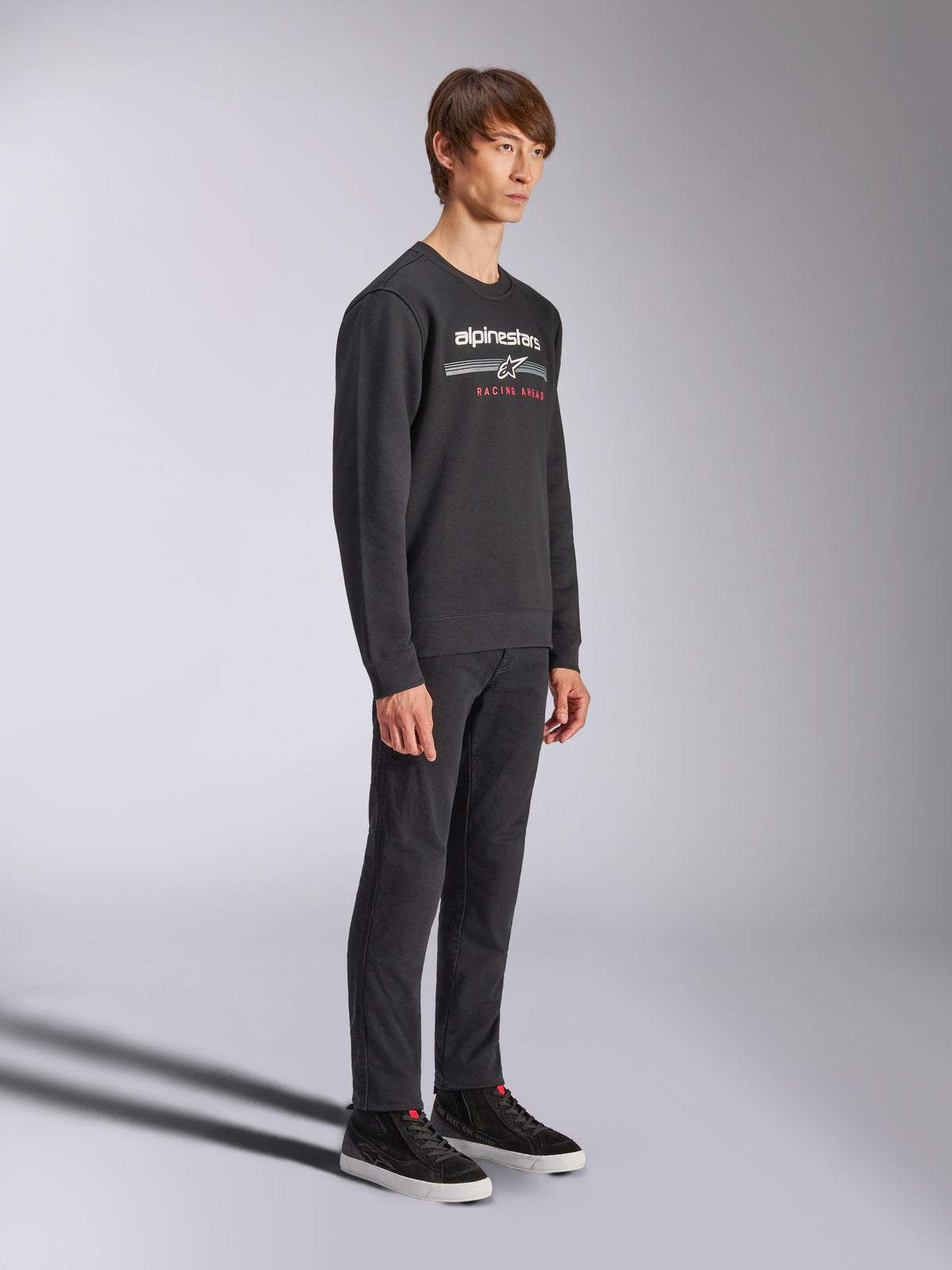 Sweatshirt Alpinestars Bettern Crew, pull à col rond noir, logo Alpinestars blanc avec l'inscription 'RACING AHEAD' en rouge sur la poitrine, associé à un pantalon noir et des baskets, style streetwear décontracté inspiré du sport automobile