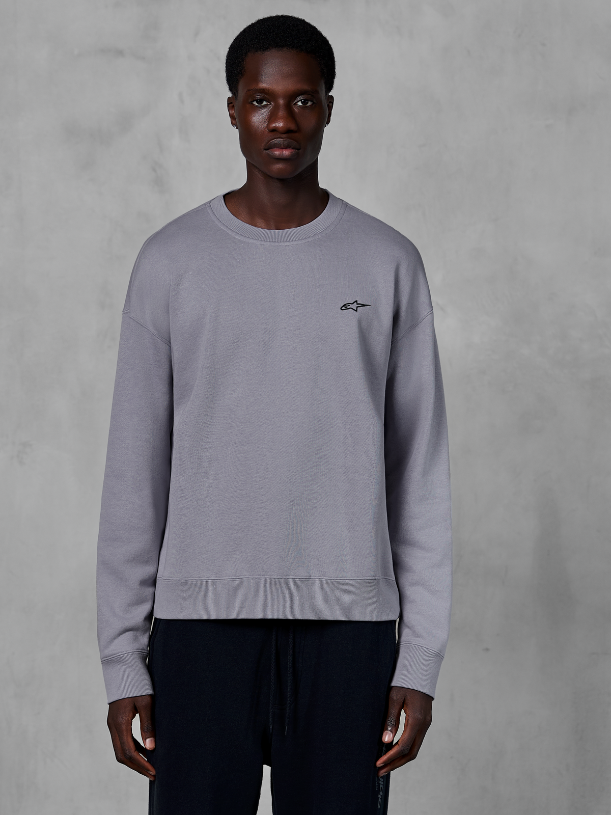 Alpinestars Essentials Ageless Crew, sweat-shirt à col rond argenté porté par un mannequin, avec un petit logo étoile noir sur la poitrine, coupe décontractée avec poignets et ourlet côtelés, style streetwear décontracté inspiré du sport automobile