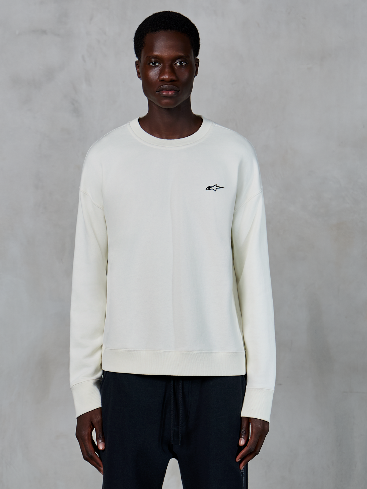 Alpinestars Essentials Ageless Crew, sweat-shirt à col rond blanc cassé, petit logo Astars noir sur la poitrine gauche, vêtement sportswear décontracté, texture polaire premium, porté par un mannequin avec un jogging noir