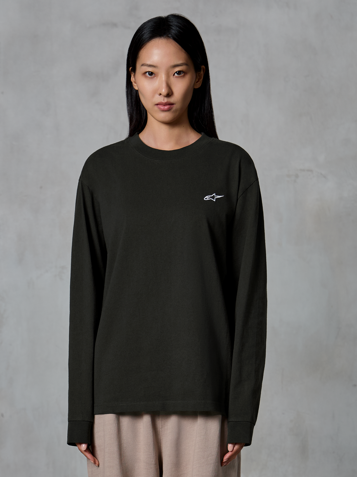 Modèle portant le t-shirt à manches longues Alpinestars Essentials Ageless, Off Black, avec un petit logo Alpinestars blanc sur la poitrine, associé à un pantalon beige, style streetwear décontracté