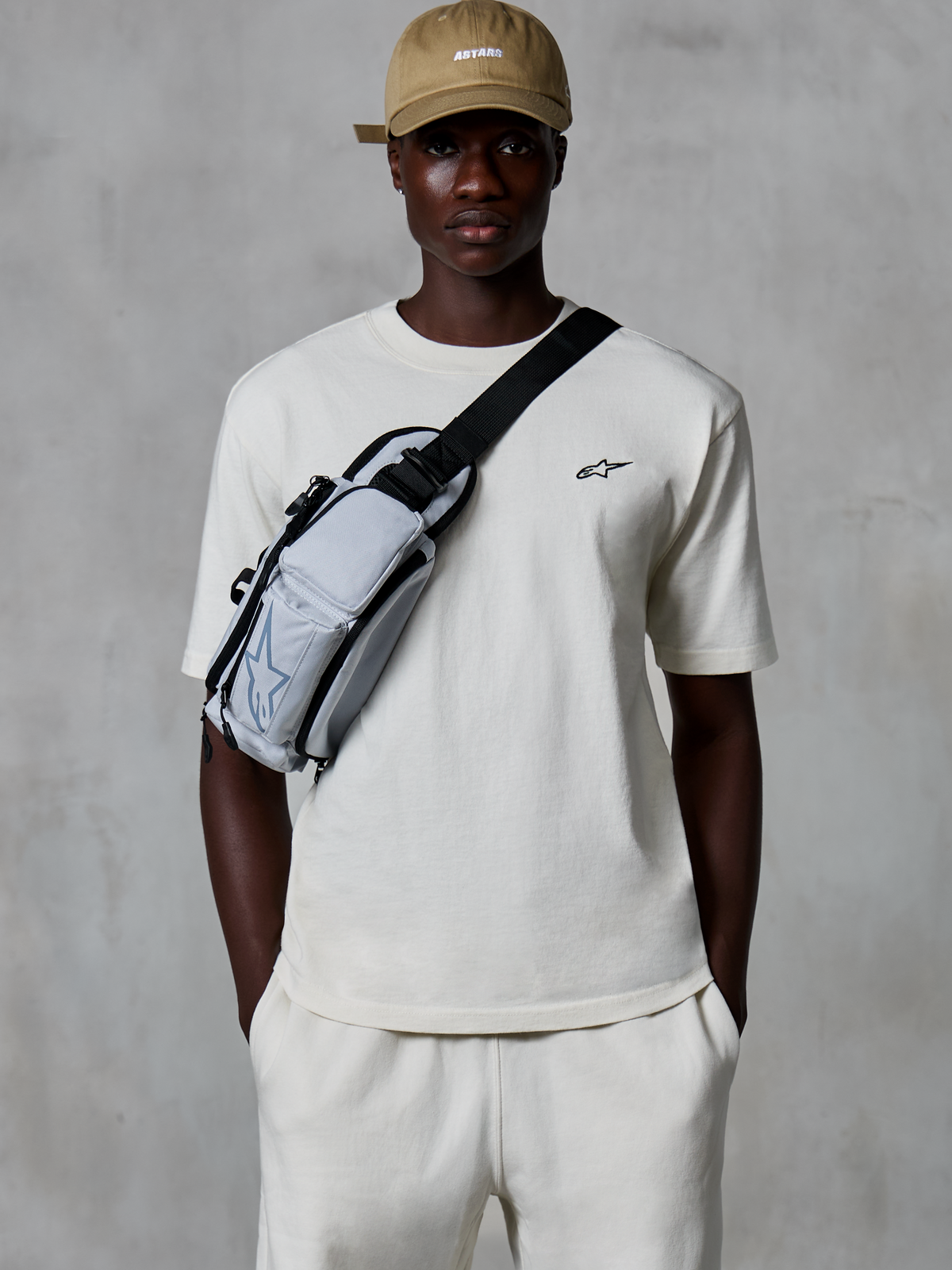 Alpinestars Essentials Ageless SS Tee, t-shirt à manches courtes, blanc cassé, porté avec un short assorti et une casquette beige, arborant un petit logo noir sur la poitrine et un sac bandoulière gris, streetwear décontracté inspiré des sports mécaniques