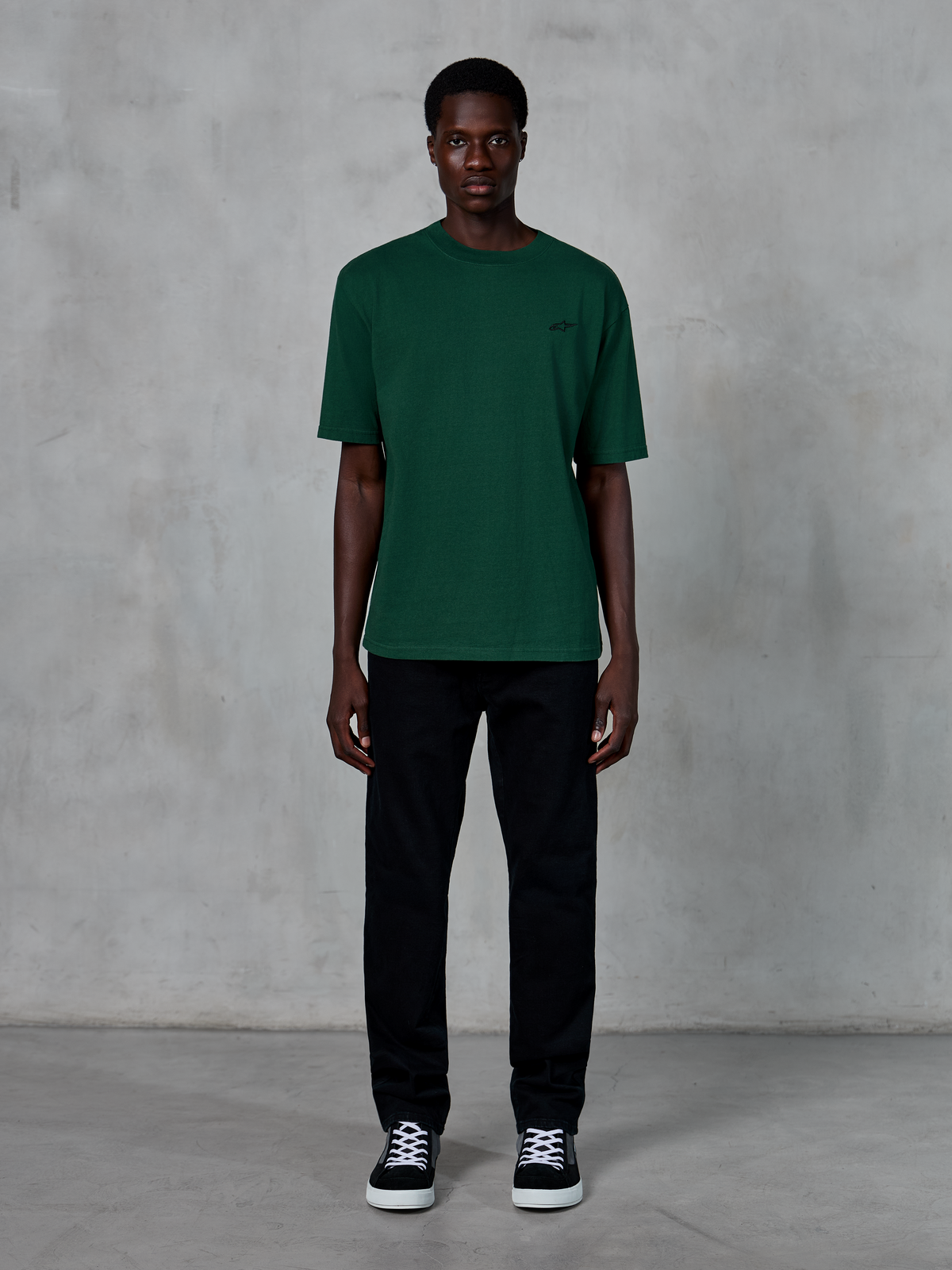T-shirt Alpinestars Essentials Ageless SS, t-shirt à manches courtes, vert sapin, avec un logo noir discret sur la poitrine, porté par un mannequin avec un pantalon noir et des baskets, style streetwear décontracté avec une coupe décontractée