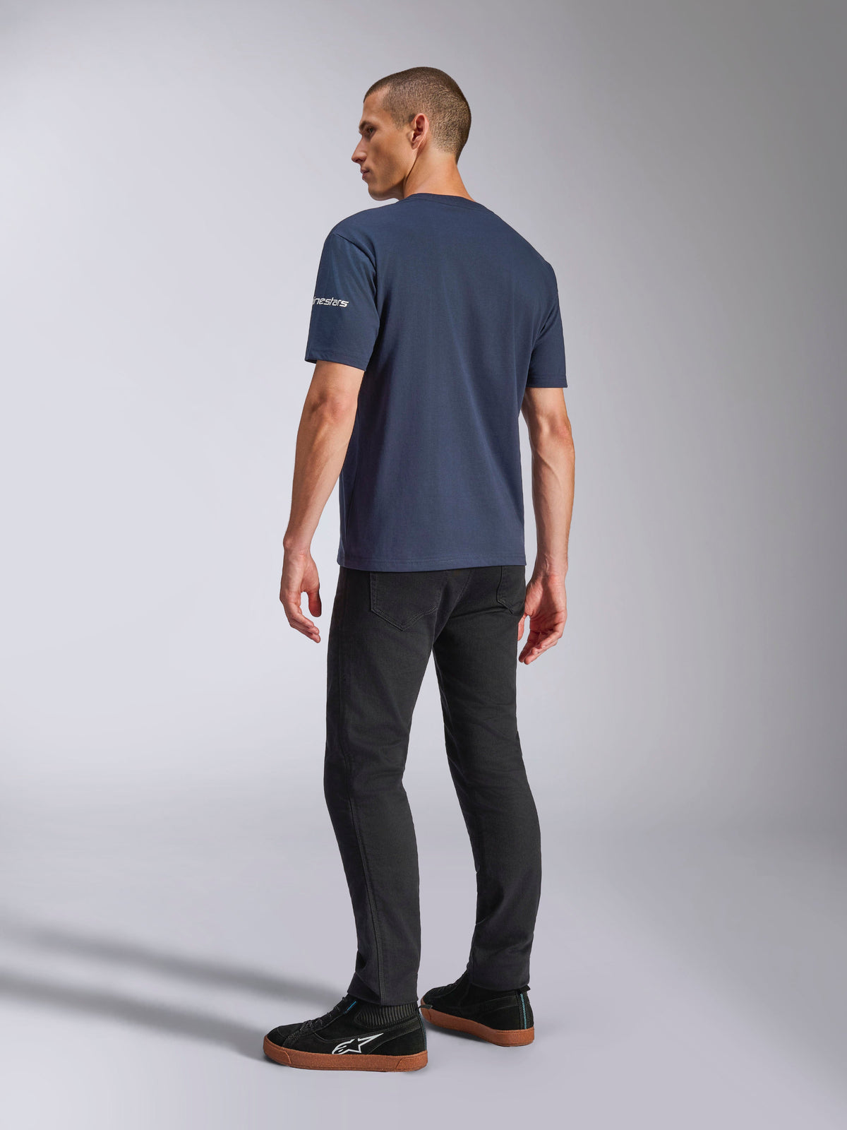 Alpinestars Frontal CSF Tee, t-shirt à manches courtes, bleu marine, vue de dos sur mannequin montrant le logo sur la manche, porté avec un pantalon noir et des baskets, streetwear décontracté inspiré du sport automobile