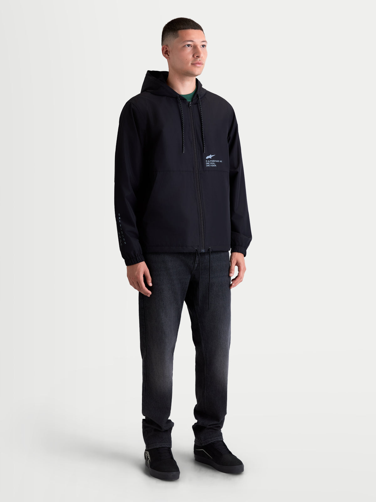 Alpinestars Speeder Resolve, veste coupe-vent de sportswear pour homme, noire, avec capuche à cordon de serrage réglable et logos d'inspiration motorsport sur la poitrine et la manche, portée par un modèle pour un look urbain