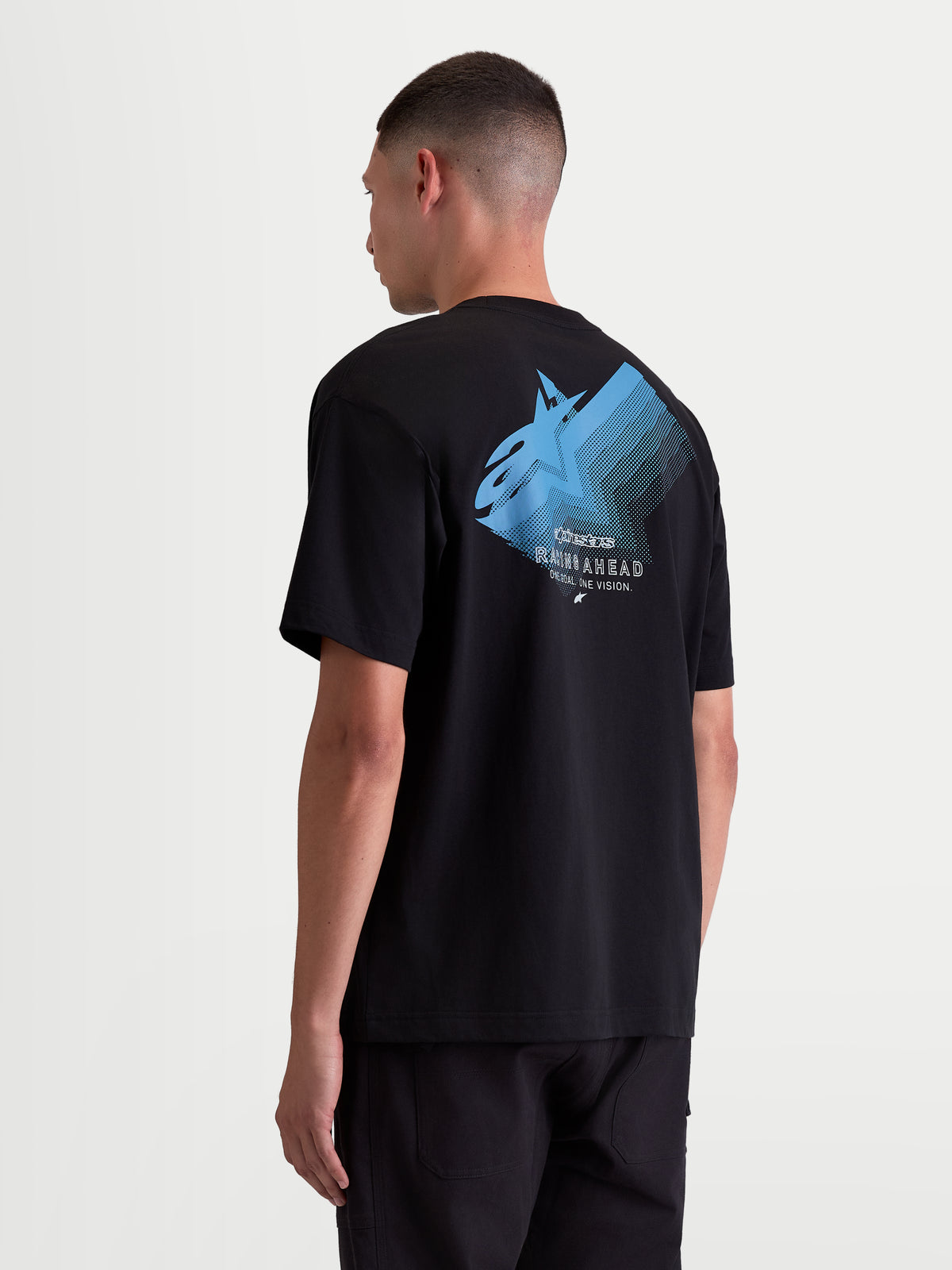 Alpinestars Ghost Oversized CSF, t-shirt de sportswear à manches courtes pour homme, noir avec graphique bleu. Vue de dos sur modèle montrant un grand logo Astars en dégradé avec les textes 'Racing Ahead' et 'One Goal. One Vision.'. Coupe oversized décontractée idéale pour un style après-moto casual.