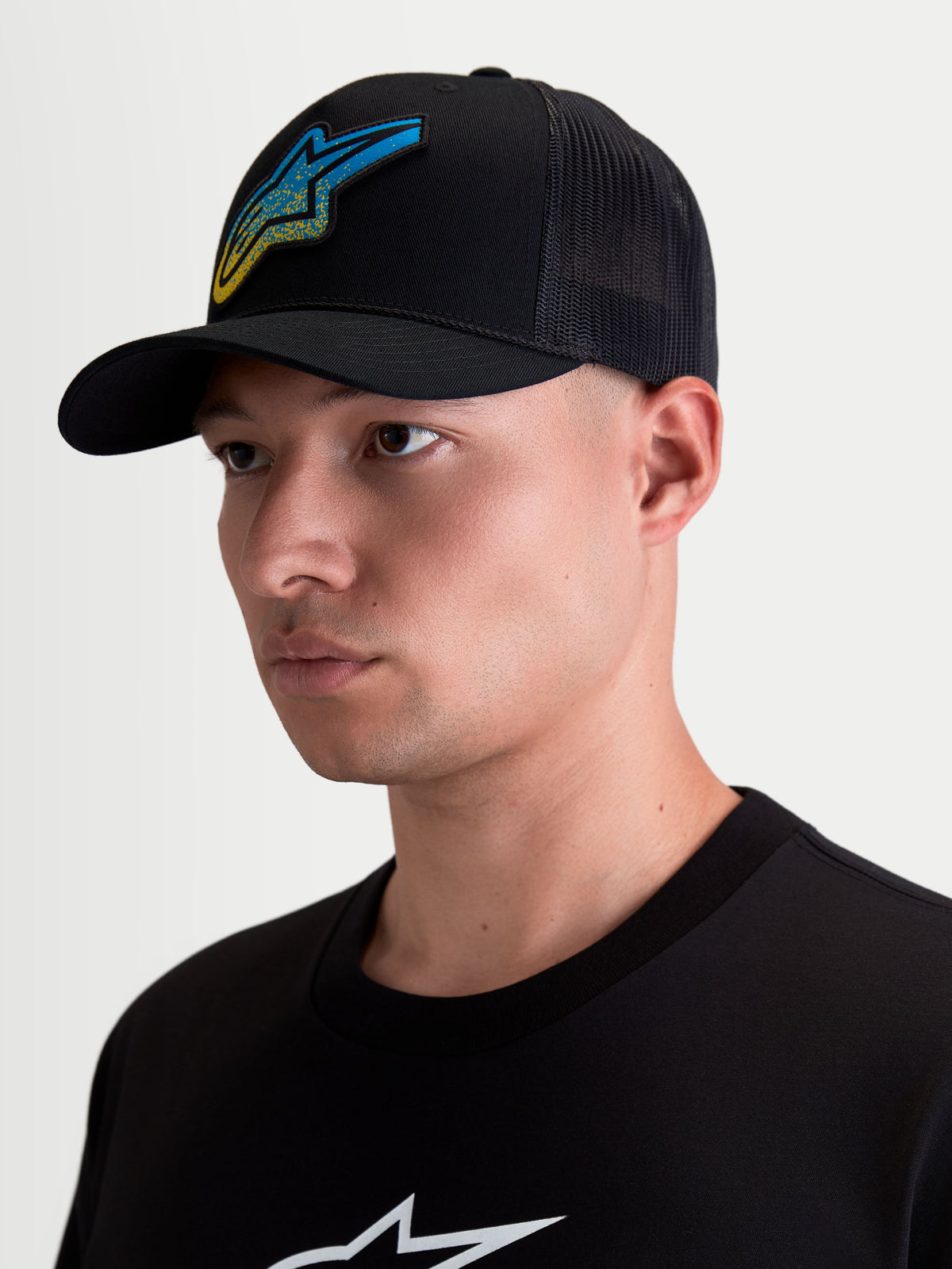 Alpinestars Casquette Trucker Meteorite, casquette de style sportswear pour homme, noire avec un écusson tissé Astars bleu et jaune à texture gravier, visière incurvée et arrière en maille respirante, portée par un modèle.