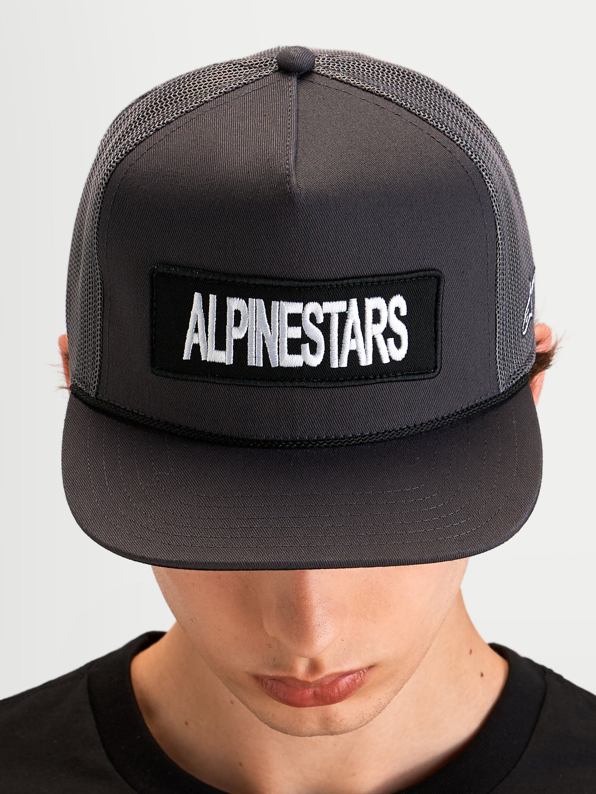 Casquette Trucker Ten d'Alpinestars, casquette sportswear à visière plate portée par un modèle, gris foncé, avec un patch logo Alpinestars à l'avant et une couronne en mesh structurée