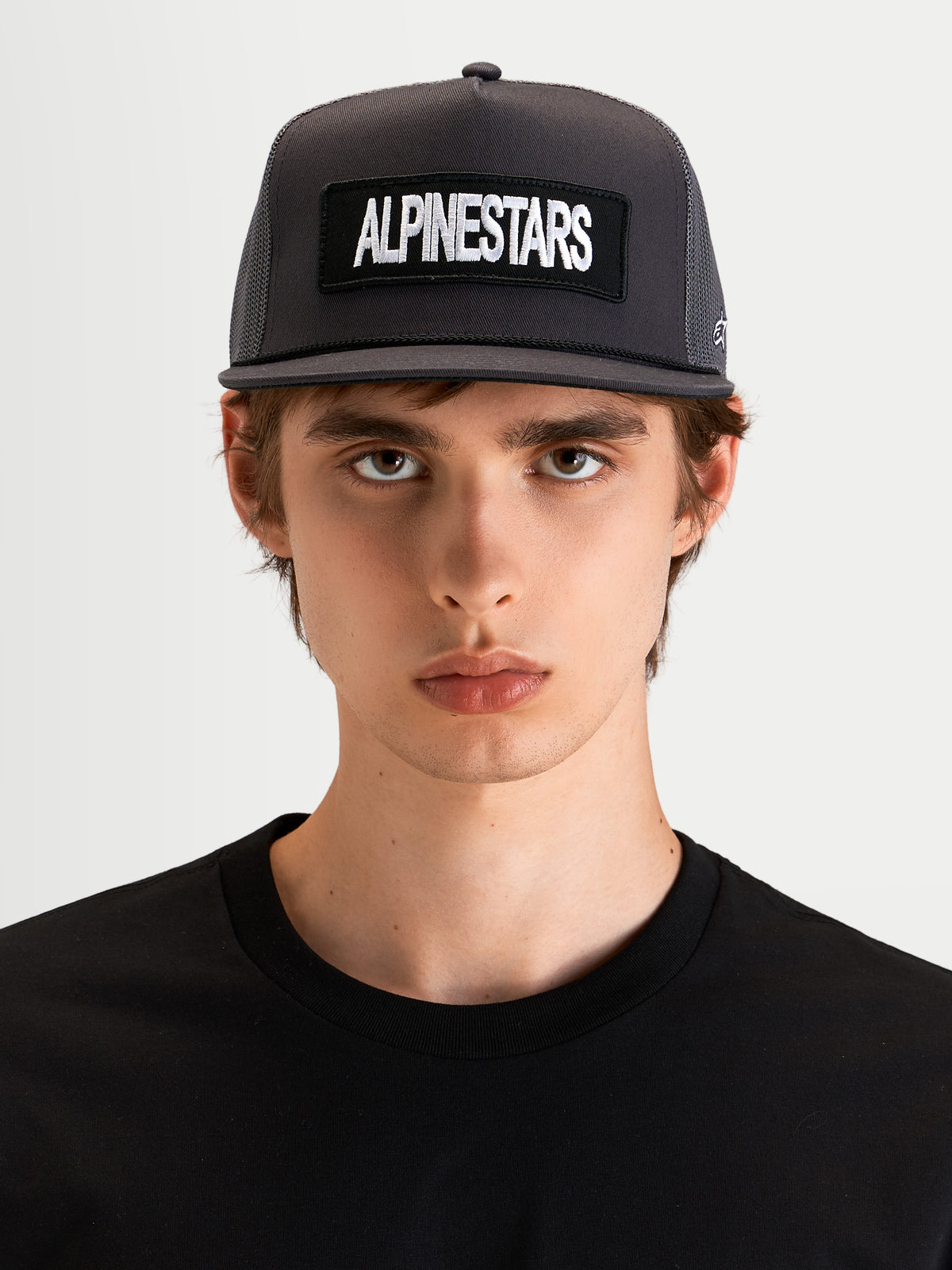 Alpinestars Casquette Trucker Ten, casquette sportswear à visière plate gris foncé, portée par un modèle, avec couronne structurée et écusson brodé du logo Alpinestars à l'avant