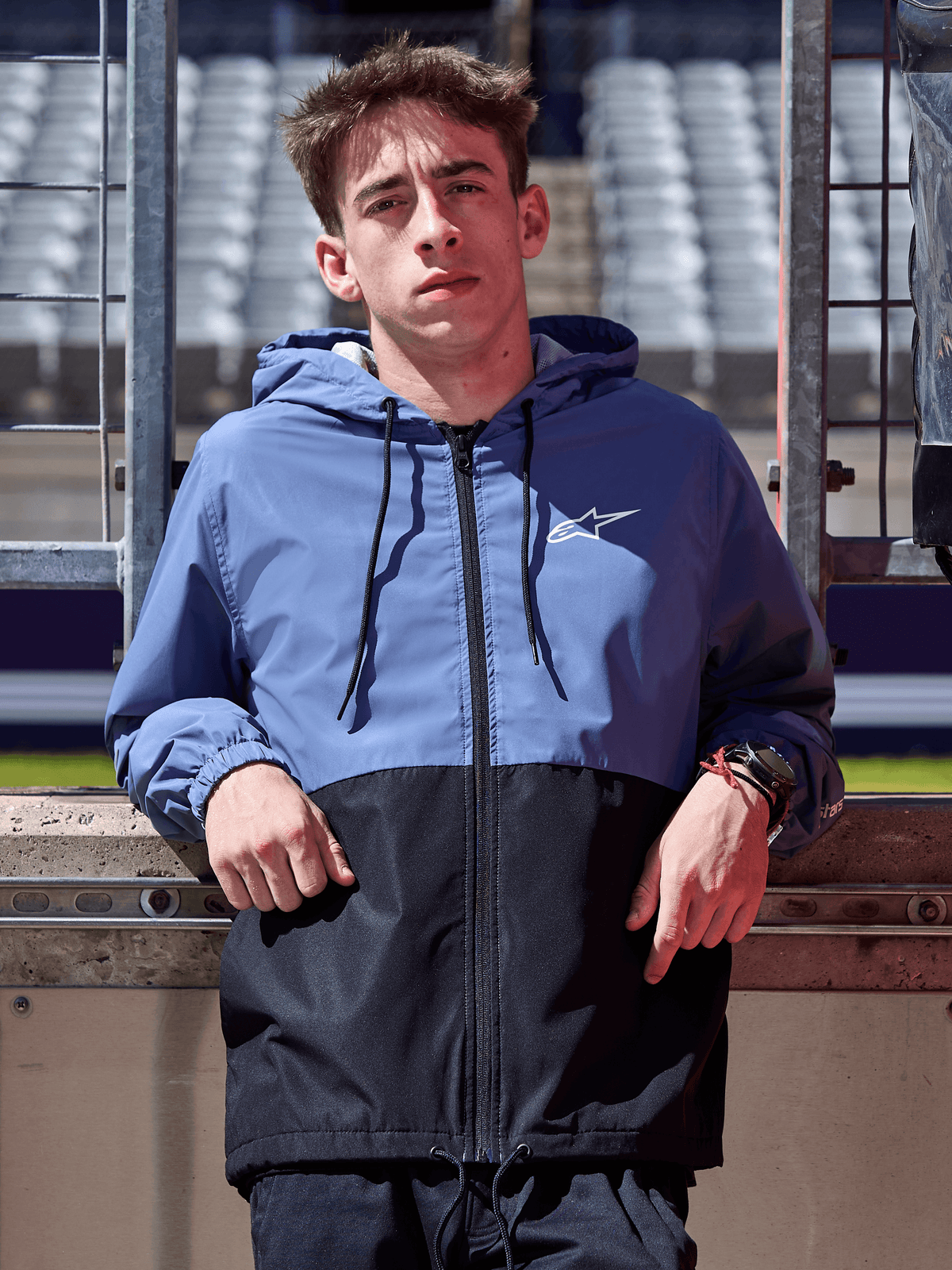 Veste coupe-vent Alpinestars Speeder, bleu ardoise et noir, coupe-vent à capuche avec design bicolore, logo blanc sur la poitrine, vêtement lifestyle décontracté porté sur un circuit de course