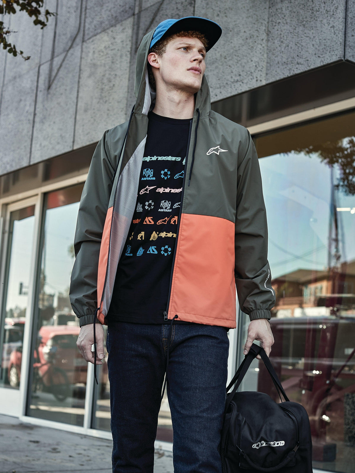 Homme portant la veste coupe-vent Alpinestars Speeder en Military Orange, avec un design color-block vert militaire et orange, à capuche avec doublure en mesh, associée à un t-shirt noir à logos graphiques, un jean bleu et un sac de sport noir Alpinestars, look lifestyle sportswear décontracté
