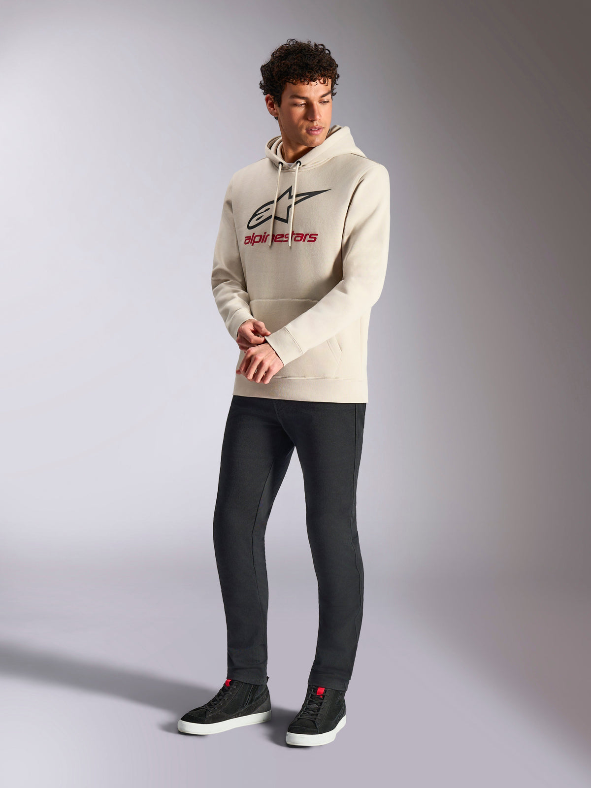 Sweat à capuche Alpinestars Always V3, hoodie en polaire blanc cassé, logo linéaire à l'avant avec étoile noire et marquage rouge, porté par un mannequin homme avec un pantalon noir et des baskets noires, style streetwear décontracté motorsport