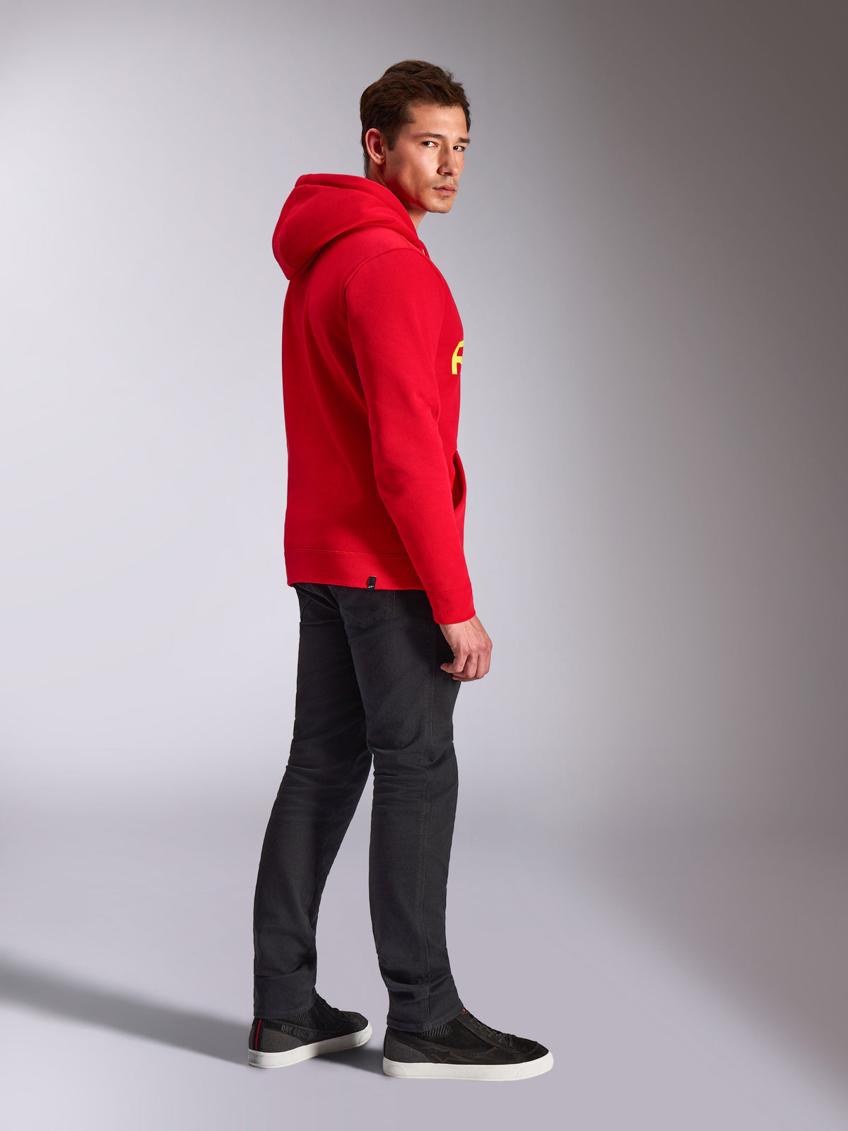 Alpinestars Sweat à Capuche Linear Wordmark V3, rouge, streetwear décontracté, modèle vu de profil et de dos portant un sweat à capuche rouge avec logo jaune, jean gris foncé et baskets noires, photo lifestyle en studio
