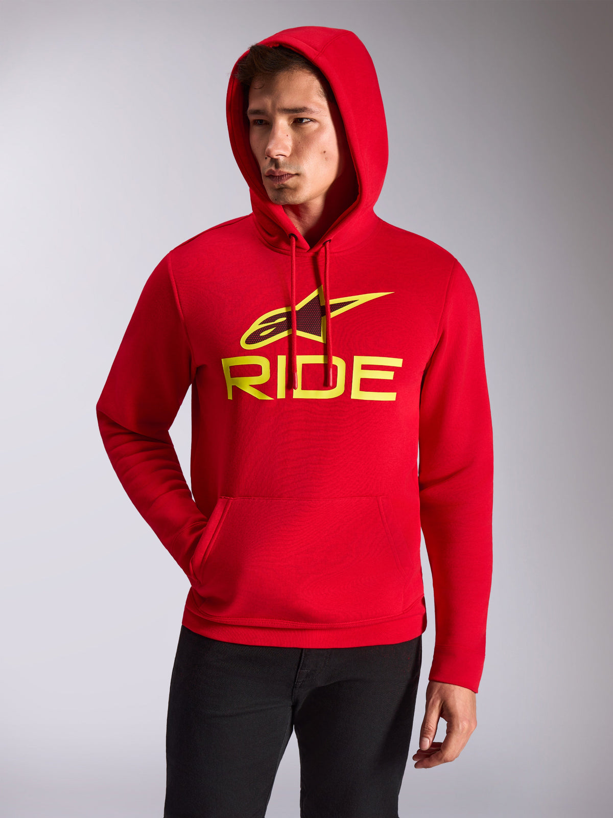 Sweat à capuche Alpinestars Ride V3, modèle masculin portant un sweat à capuche en polaire rouge, logo RIDE jaune haute visibilité et étoile Alpinestars sur la poitrine, style streetwear décontracté, poche kangourou à l'avant, porté avec un pantalon noir.