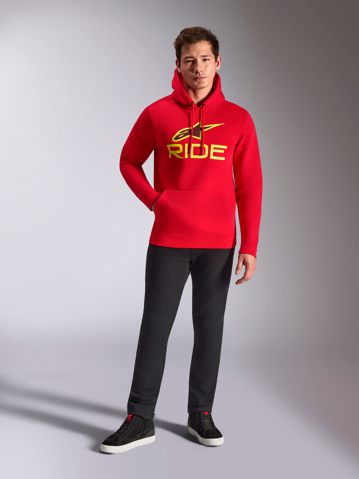 Sweat à capuche Alpinestars Ride V3, polaire rouge, logo RIDE jaune vif avec symbole Astars sur la poitrine, porté avec un pantalon noir et des baskets montantes noires, streetwear décontracté inspiré du sport automobile