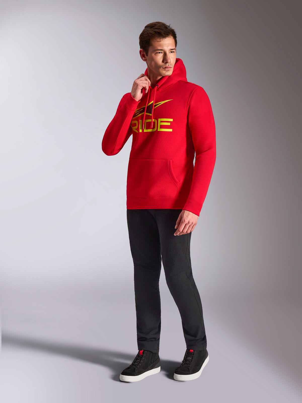 Sweat à capuche Alpinestars Ride V3, sweat-shirt rouge, logo 'RIDE' jaune et noir sur la poitrine, porté par un mannequin avec un pantalon noir et des baskets noires, style streetwear décontracté inspiré du sport automobile