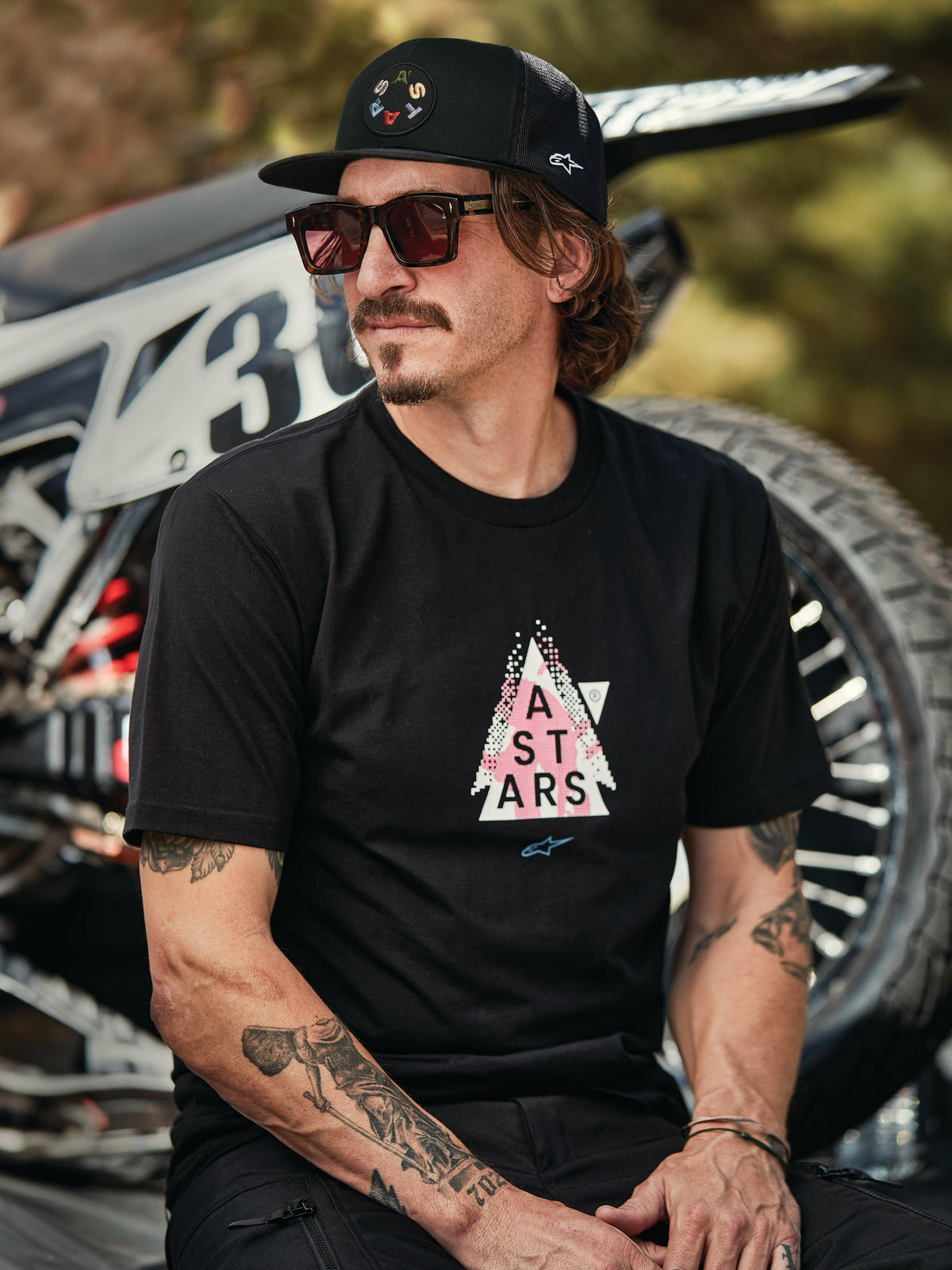 Casquette Trucker Alpinestars Gambit en noir avec écusson logo circulaire, portée avec le t-shirt Alpinestars Soloist SS noir, photo lifestyle mettant en scène un homme avec des lunettes de soleil et des tatouages assis devant une moto de motocross.