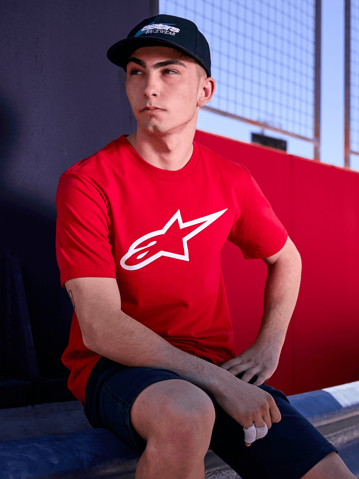 Casquette Alpinestars Profile noire, photo lifestyle d'un homme portant la casquette à visière courbée avec un t-shirt rouge arborant un logo Alpinestars blanc, style streetwear décontracté inspiré du sport automobile