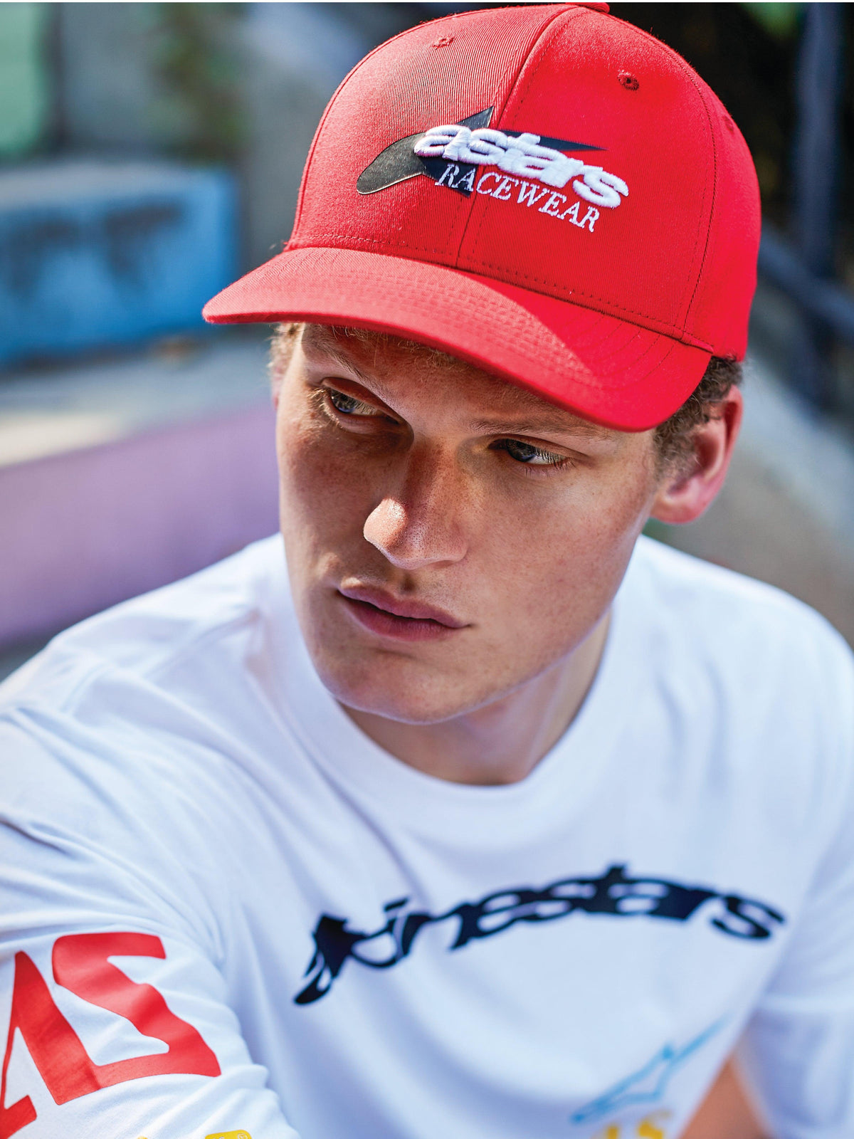 Homme portant une casquette Alpinestars Profile, snapback rouge à visière courbée avec broderie frontale « astars RACEWEAR » blanche et noire, associée à un t-shirt graphique blanc à manches longues Alpinestars, style décontracté motorsport
