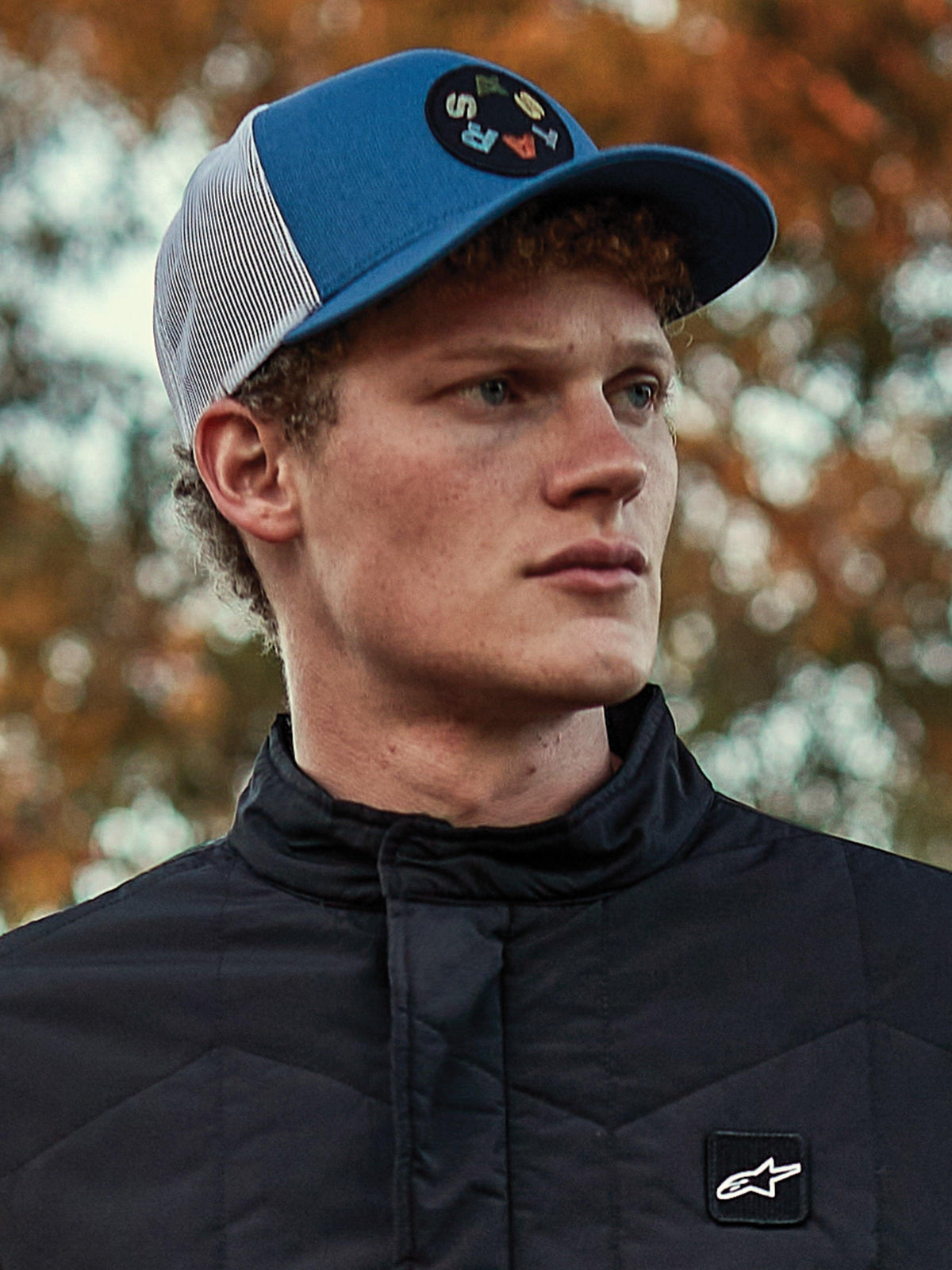 Homme portant une casquette Alpinestars Gambit Trucker, casquette bleue et blanche avec dos en maille et écusson logo Astars circulaire, associée à une veste matelassée noire Alpinestars, portrait lifestyle décontracté inspiré du sport automobile avec arrière-plan automnal