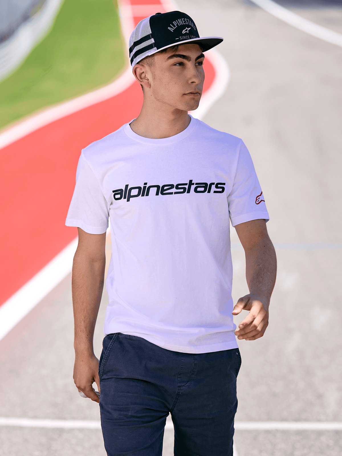 Homme portant une casquette Alpinestars Backline Trucker en noir et blanc, avec un t-shirt blanc à logo Alpinestars et un pantalon bleu foncé, style streetwear motorsport décontracté sur fond de circuit extérieur